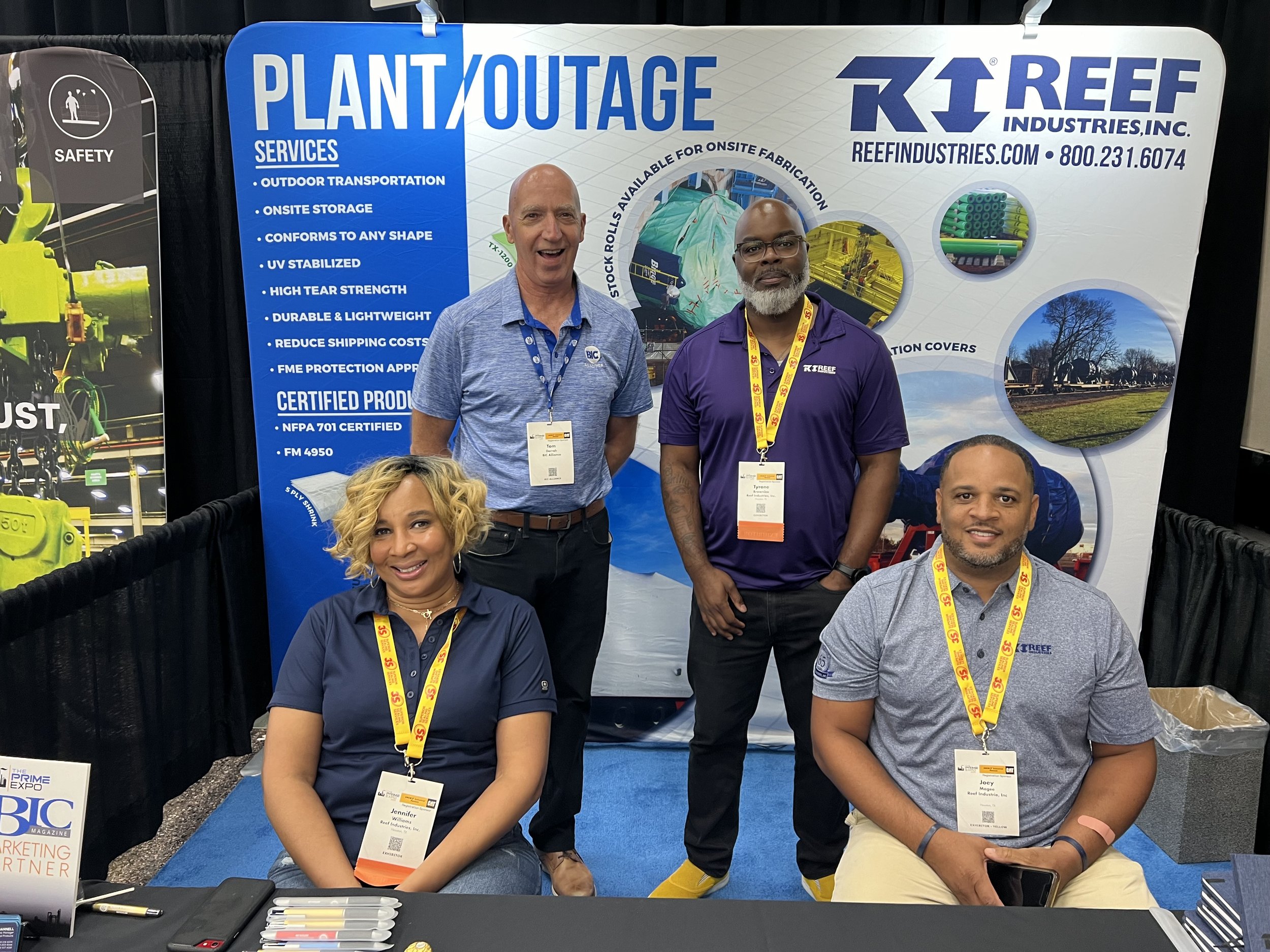 Four people posing at a trade show booth for Reef Industries, Inc., with a blue and white backdrop displaying company services and certification information. Two women are seated, and two men are standing.