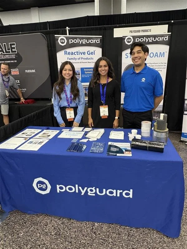 Three people standing behind a table at a trade show booth for Polyguard, displaying product brochures and samples, with black curtains and banners promoting Polyguard's Reactive Gel and Spray Foam in the background.