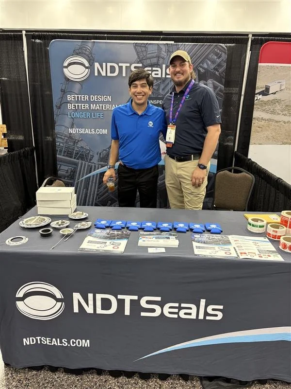 Two men standing behind a booth table at a trade show, smiling. The booth displays NDT Seals branding, with various sealing products, brochures, and promotional items on the table.