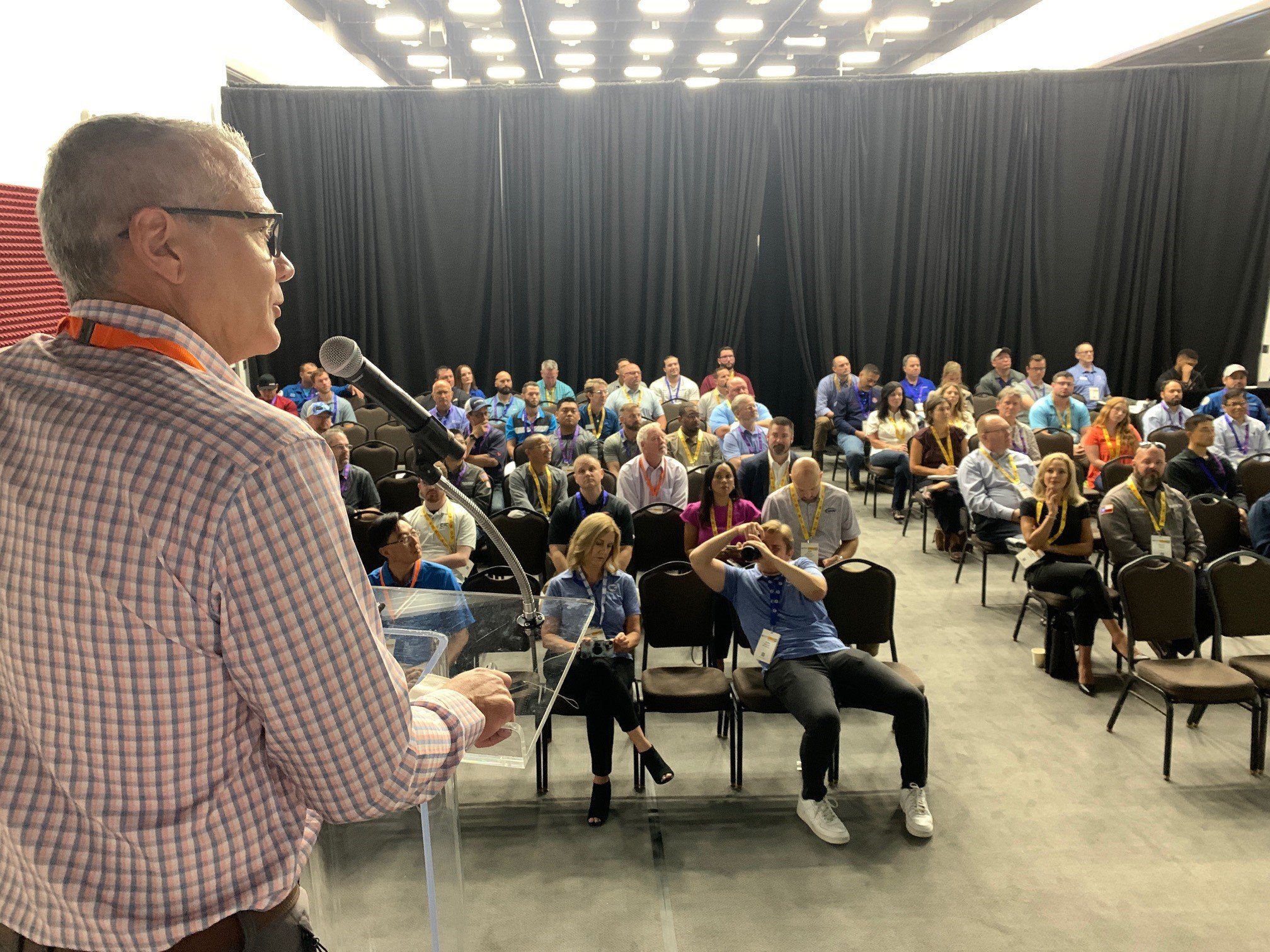 A man wearing glasses and a checkered shirt speaking at a podium with a microphone in front of an audience at a conference.