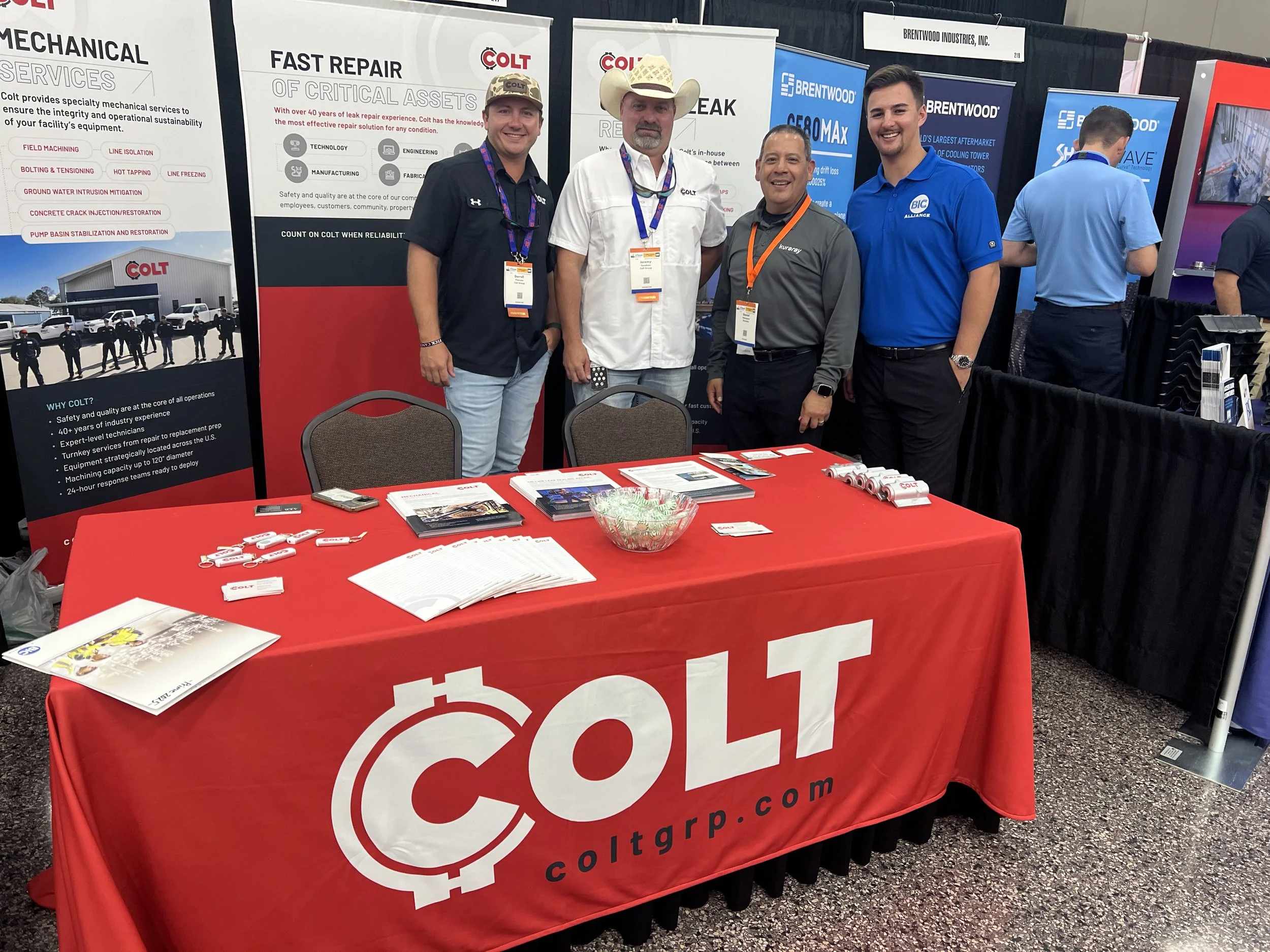 Four men standing behind a red table with brochures and white keychains, at a trade show booth for Colt. The booth has banners featuring their services and company information, with one banner showing a group photo and list of services.