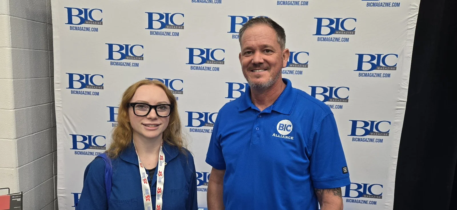 A woman and a man standing in front of a backdrop with the BIC magazine logo. The woman has long curly red hair, glasses, and a blue shirt. The man has short hair, a short beard, and is wearing a blue polo shirt with the BIC logo and the word "ALLIAN