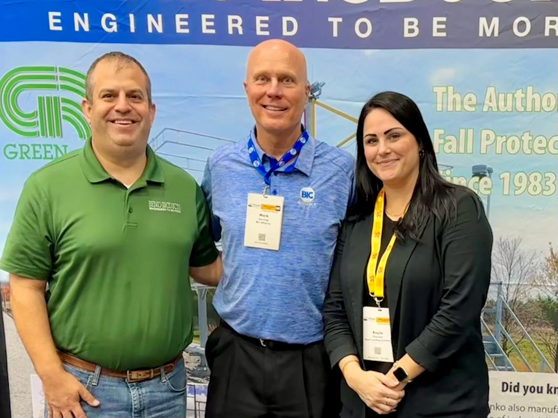 Three smiling people standing together at a conference, with a banner behind them that reads 'Engineered to be More' and includes a green logo and text about fall protection since 1983.