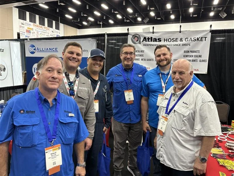 Group of six men standing at a trade show booth with banners in the background, wearing company shirts and name tags.