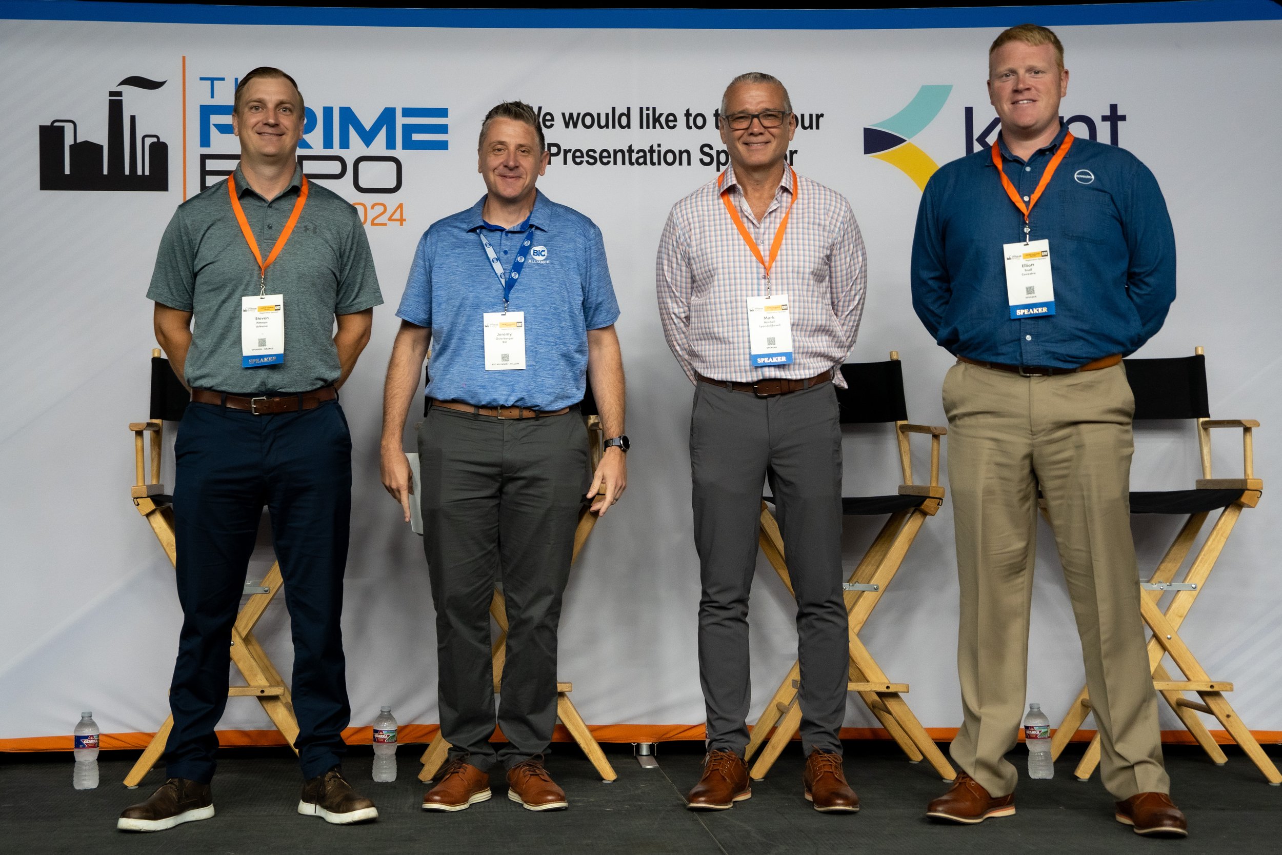 Four men standing on a stage at a conference, with chairs and water bottles on the floor behind them.