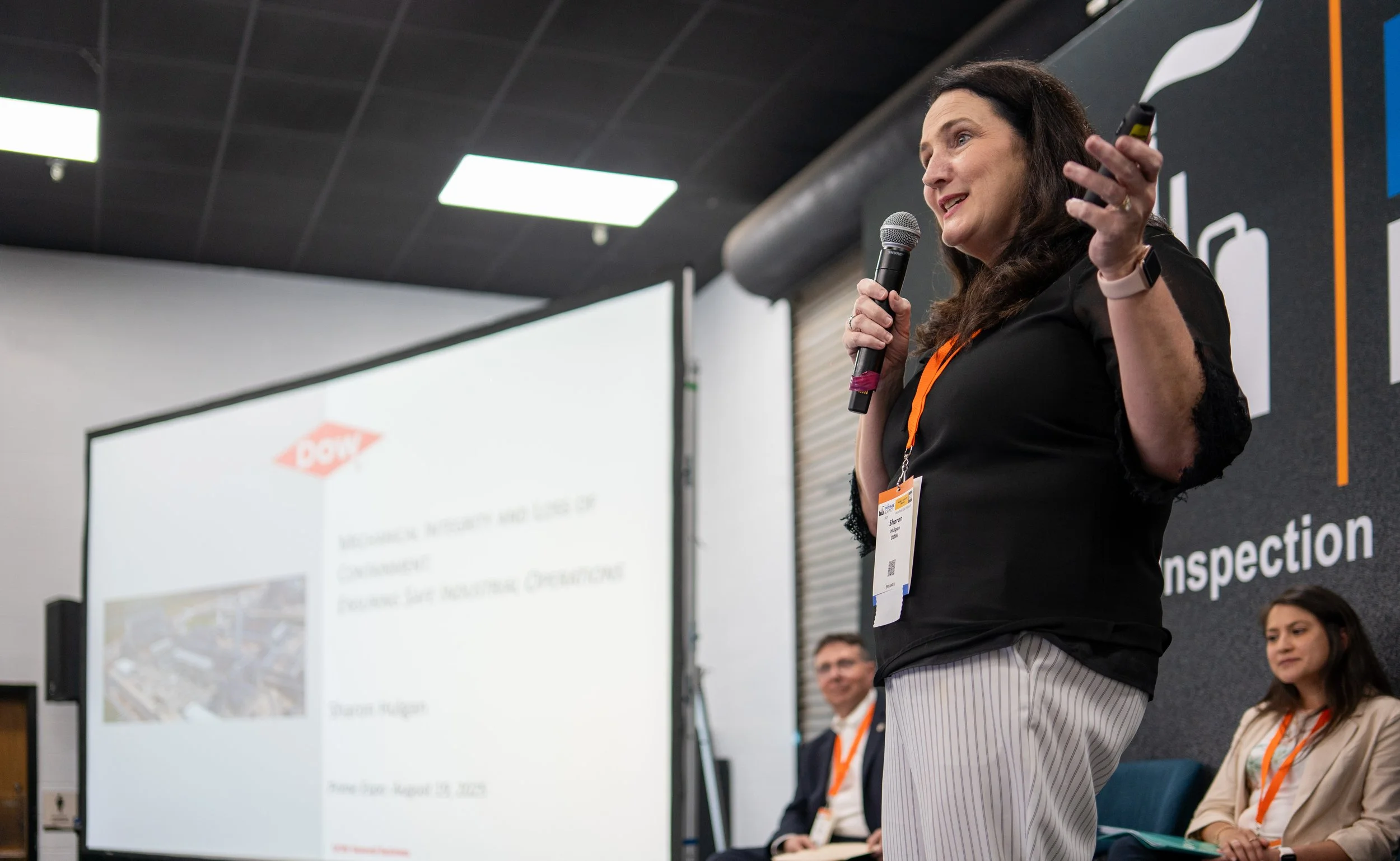 A woman speaking into a microphone during a presentation at a conference, with a projected slide behind her and other seated attendees.