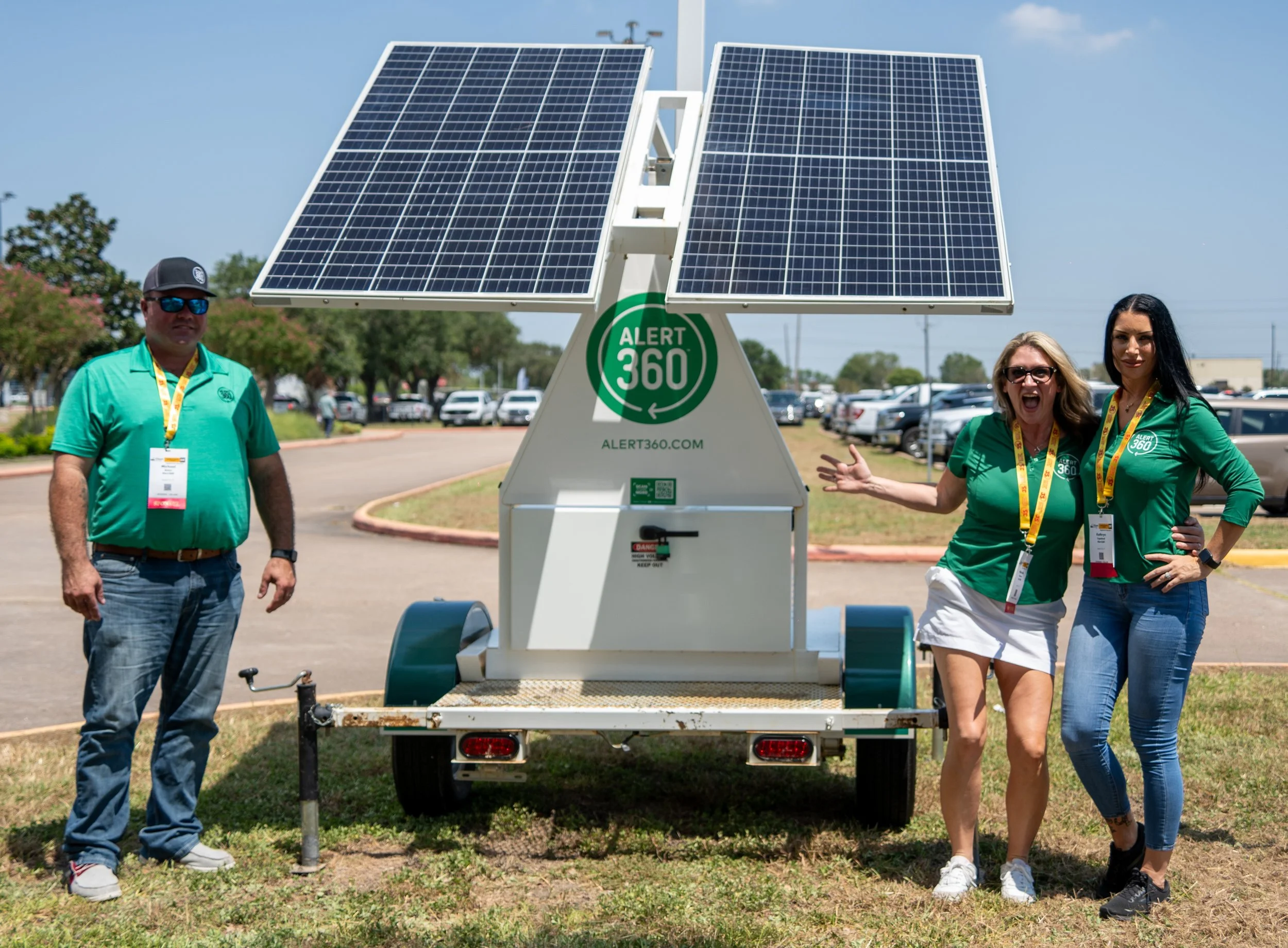 Three people standing outdoors in front of a mobile solar panel generator with the ALERT 360 logo, with a parking lot and trees in the background.