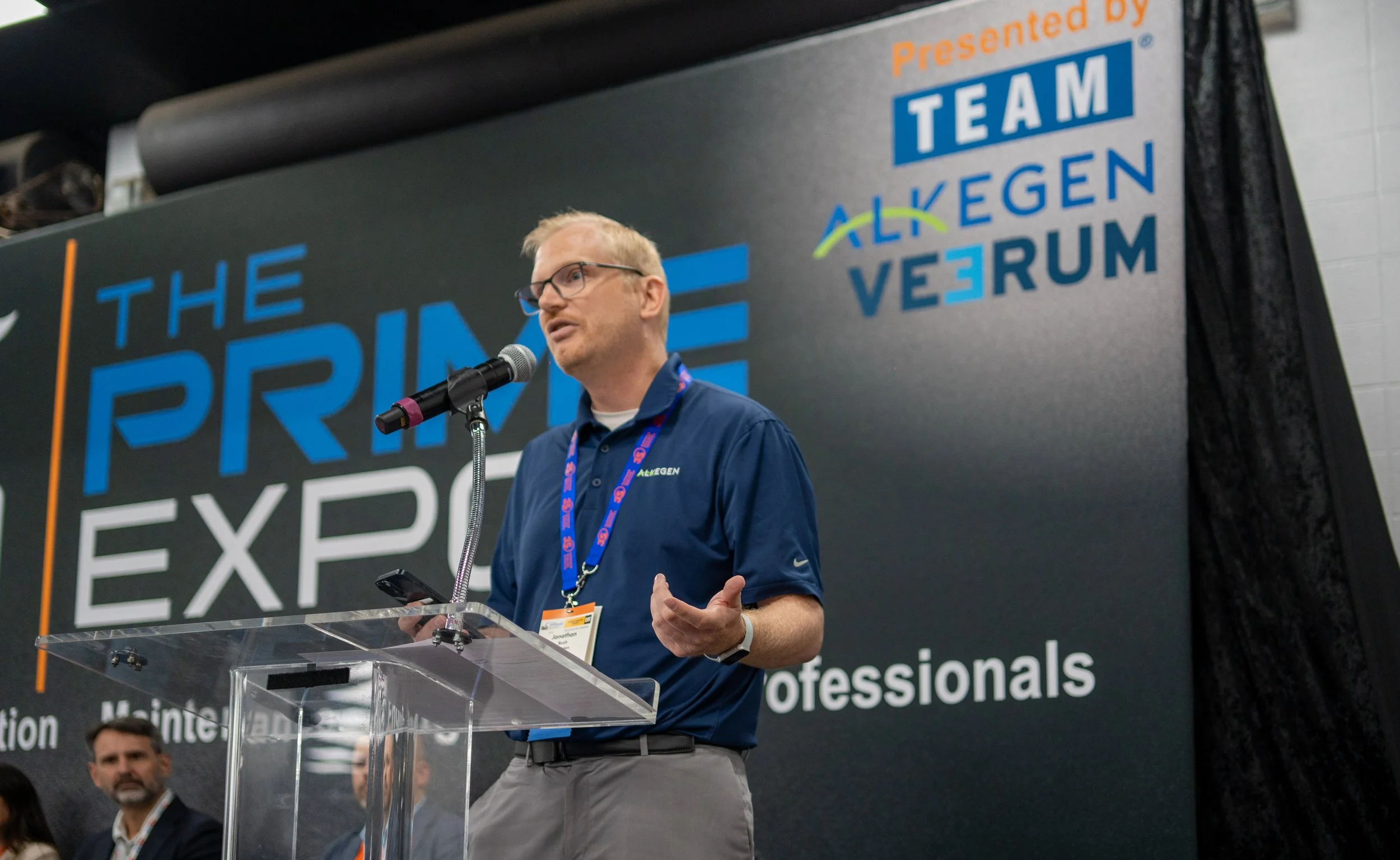 Man in blue shirt and glasses speaking at a podium during a conference, large backdrop behind him with the text 'The Prime Expo.'