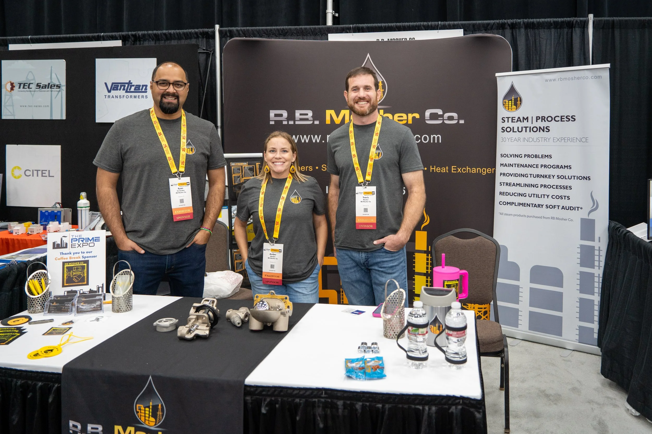 Three people standing behind a booth at a trade show, smiling at the camera. The booth displays a black banner with the text 'R.B. Mosher Co.' and a white banner with company information. The table in front has various mechanical parts, water bottles