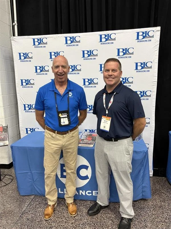 Two men standing in front of a BIC magazine backdrop at a conference. One man is wearing a blue polo shirt and khakis, the other a dark polo shirt and gray pants.