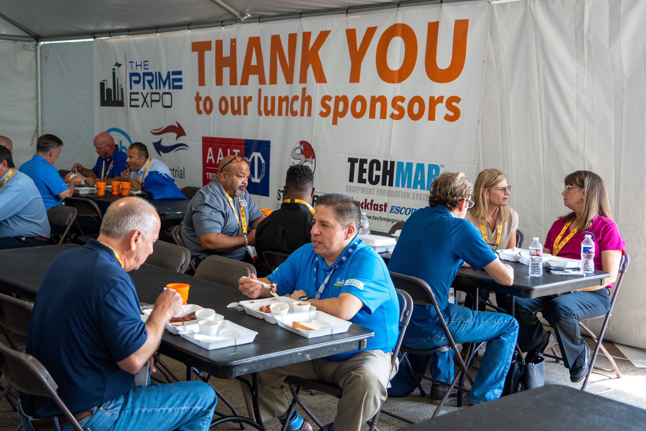 People sitting at long tables eating lunch at an expo event under a large banner that reads, 'Thank you to our lunch sponsors,' with logos of various companies.