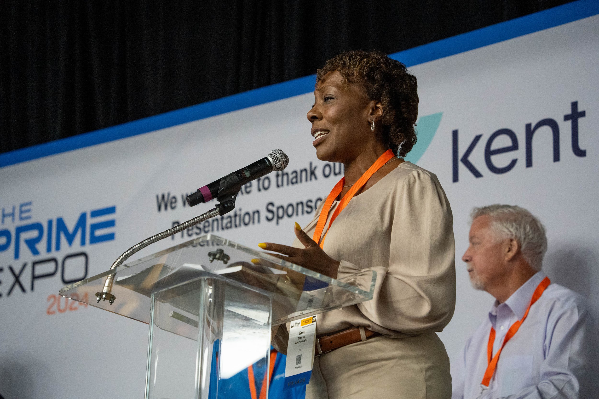 A woman speaking at a podium during an event with a sign behind her that reads "The Prime Expo 2023" and the logo "Kent." She is wearing a beige blouse, brown belt, and orange lanyard, with short curly hair, and is speaking into a microphone.