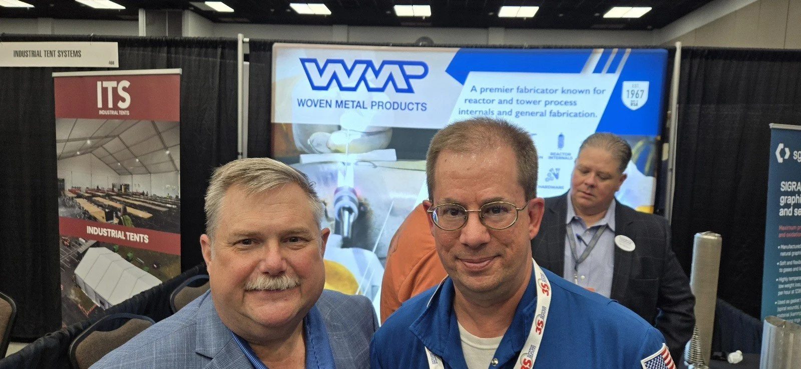Two men at an industrial trade show standing in front of a booth with banners displaying woven metal products and industrial tents.