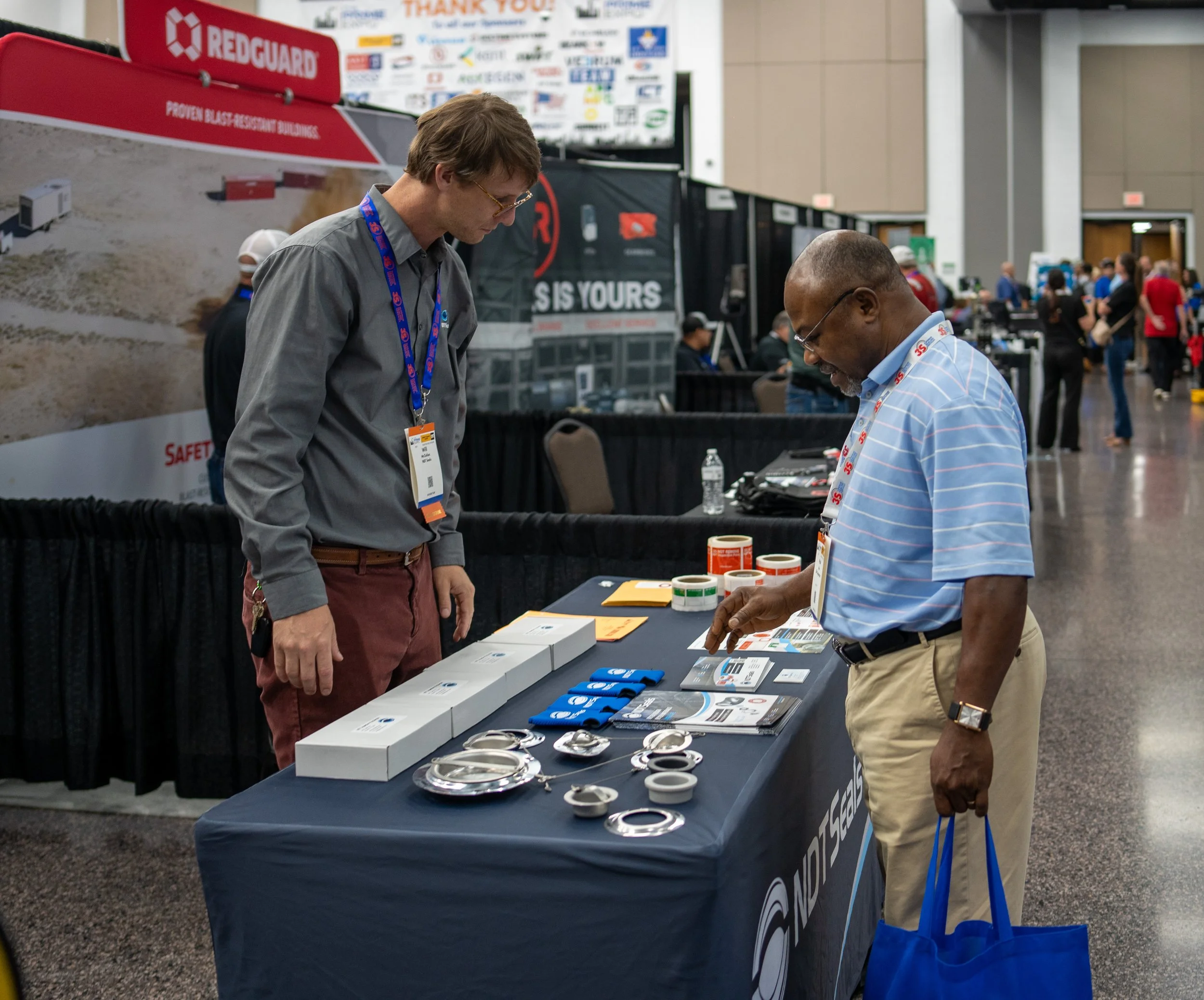 Two men at a trade show booth examining various safety equipment such as reflective gear and safety glasses. The booth has a blue tablecloth with the NPS Safety logo, and promotional materials are displayed on the table.