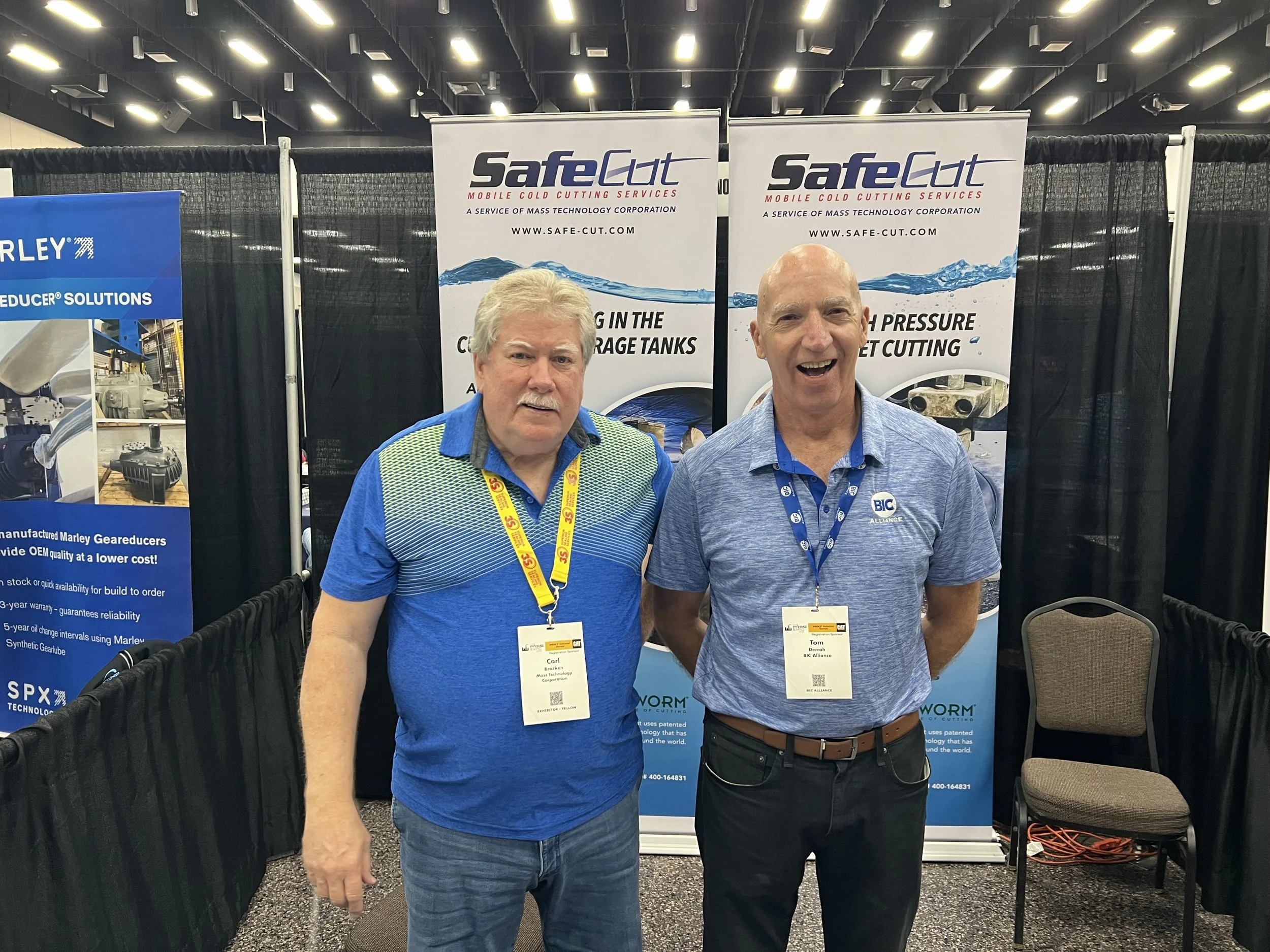Two men standing in front of trade show banners at a convention, one with grey hair and mustache wearing a blue shirt with a pattern, and the other bald with a wide smile wearing a solid blue shirt with a logo.