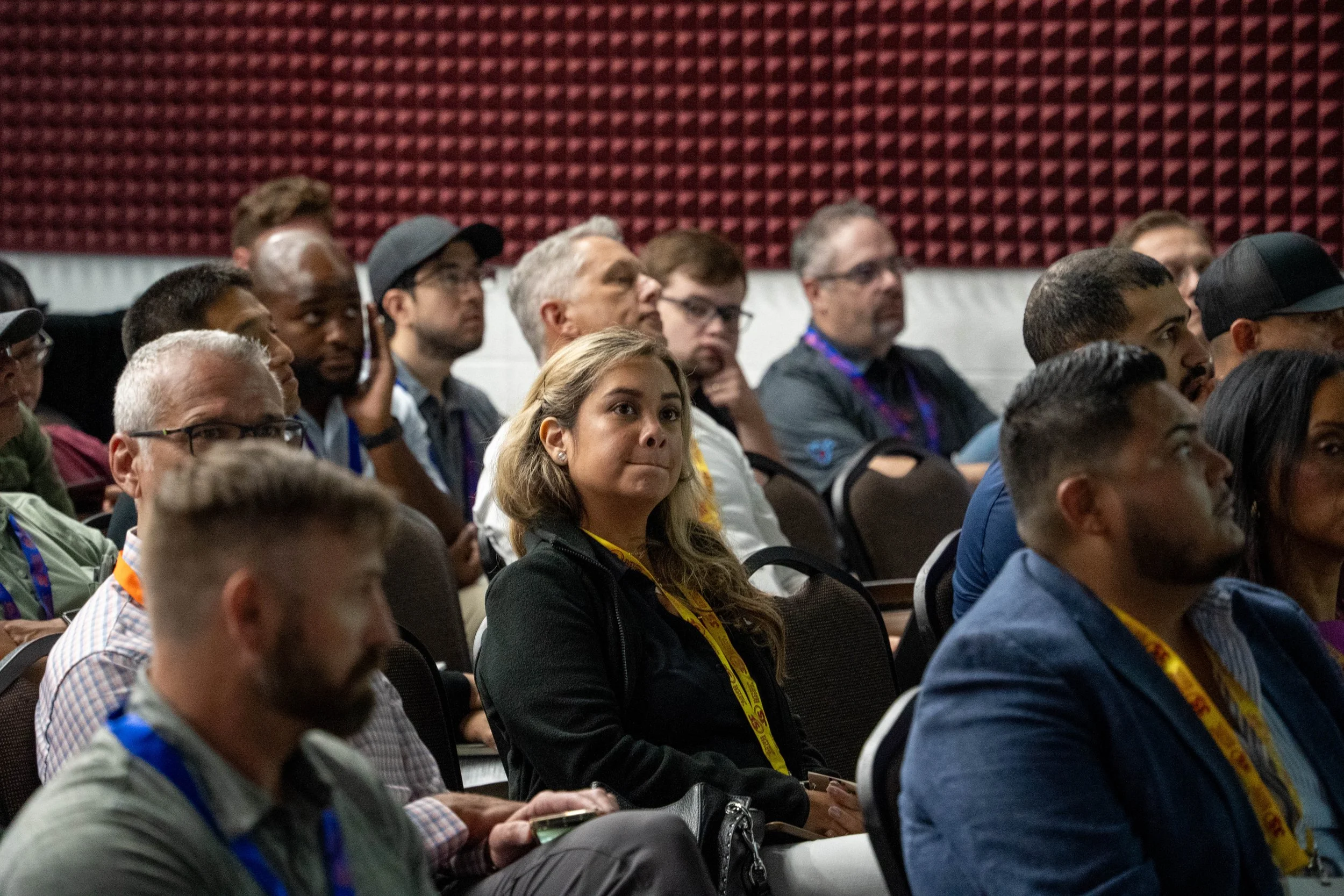 A diverse group of people attending a conference or seminar, seated and listening attentively, with some taking notes, in a room with red soundproof wall panels.