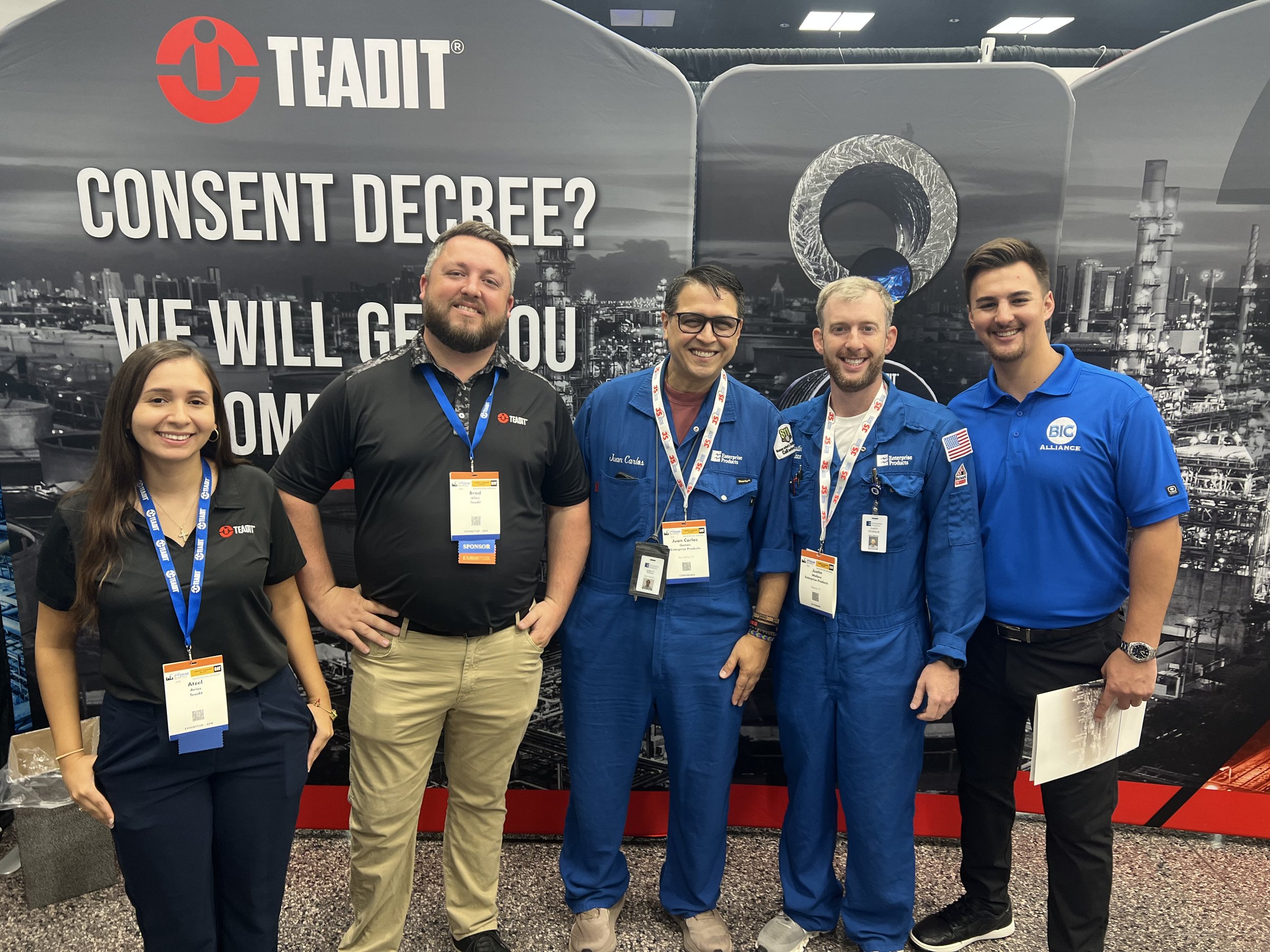 Group of five people standing together at a conference, in front of a backdrop with the Teadit logo and the words 'Consent Decree? We will get you out.' Four individuals are wearing black or blue shirts with name tags, and two are dressed in blue ast