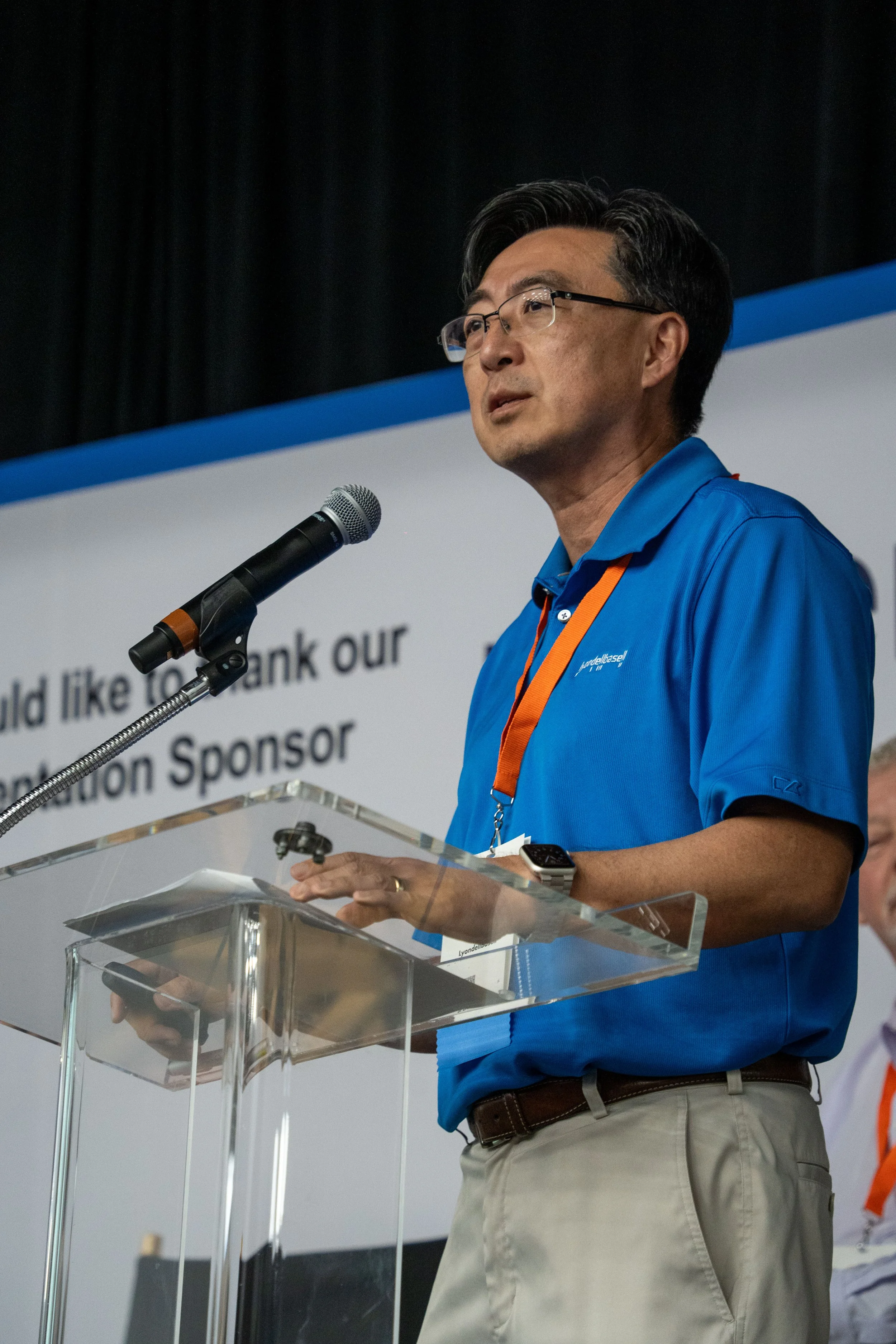 A man wearing glasses and a blue polo shirt speaking at a podium with a microphone, holding a device, during a presentation or conference.