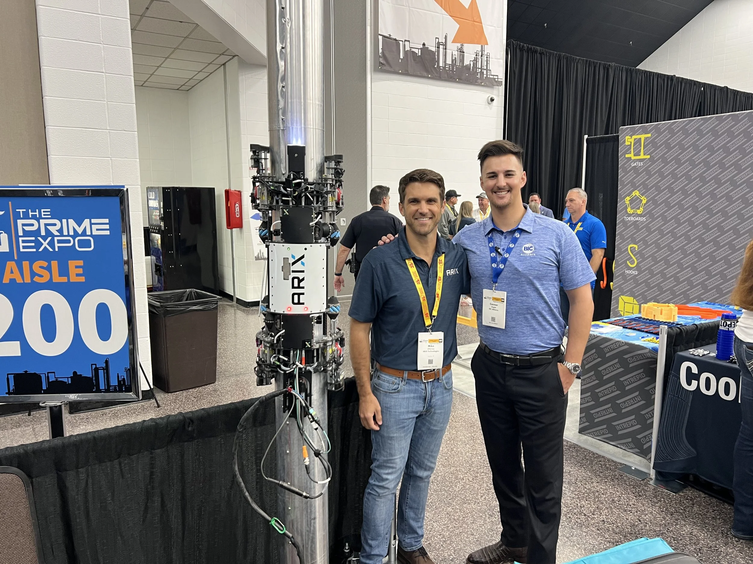Two men standing next to a robotic arm at a conference, smiling for the camera, with a sign that reads 'The Prime Expo Aisle 200' in the background.