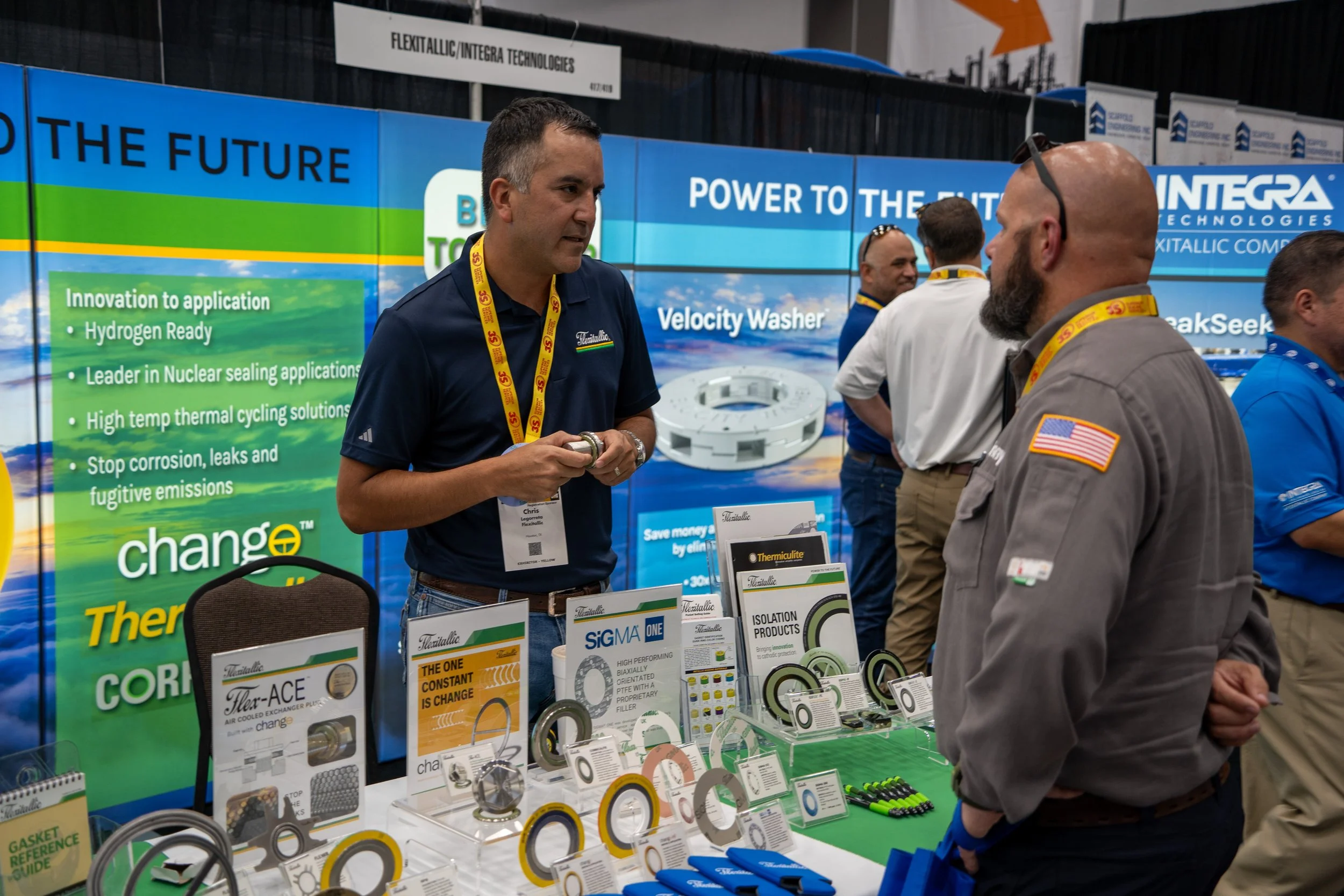 A man in a gray shirt with an American flag patch on the sleeve and a man in a navy blue polo shirt with a convention badge are talking at a trade show booth about industrial sealing products and solutions, with various product samples on display and