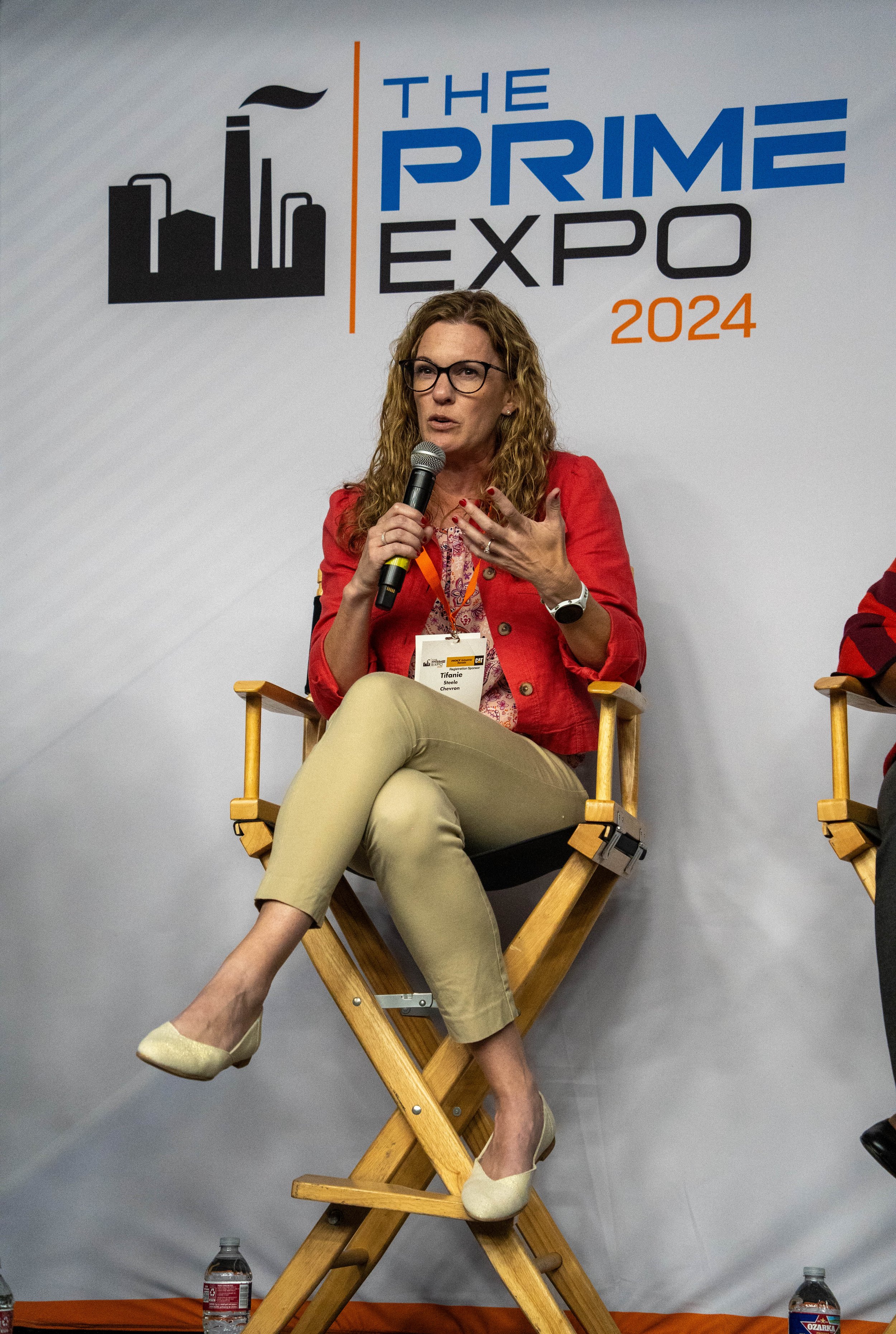 A woman with glasses, curly hair, and a red blazer speaking into a microphone at the Prime Expo 2024, seated on a wooden director's chair with water bottles on the floor, in front of a backdrop with the event logo and name.