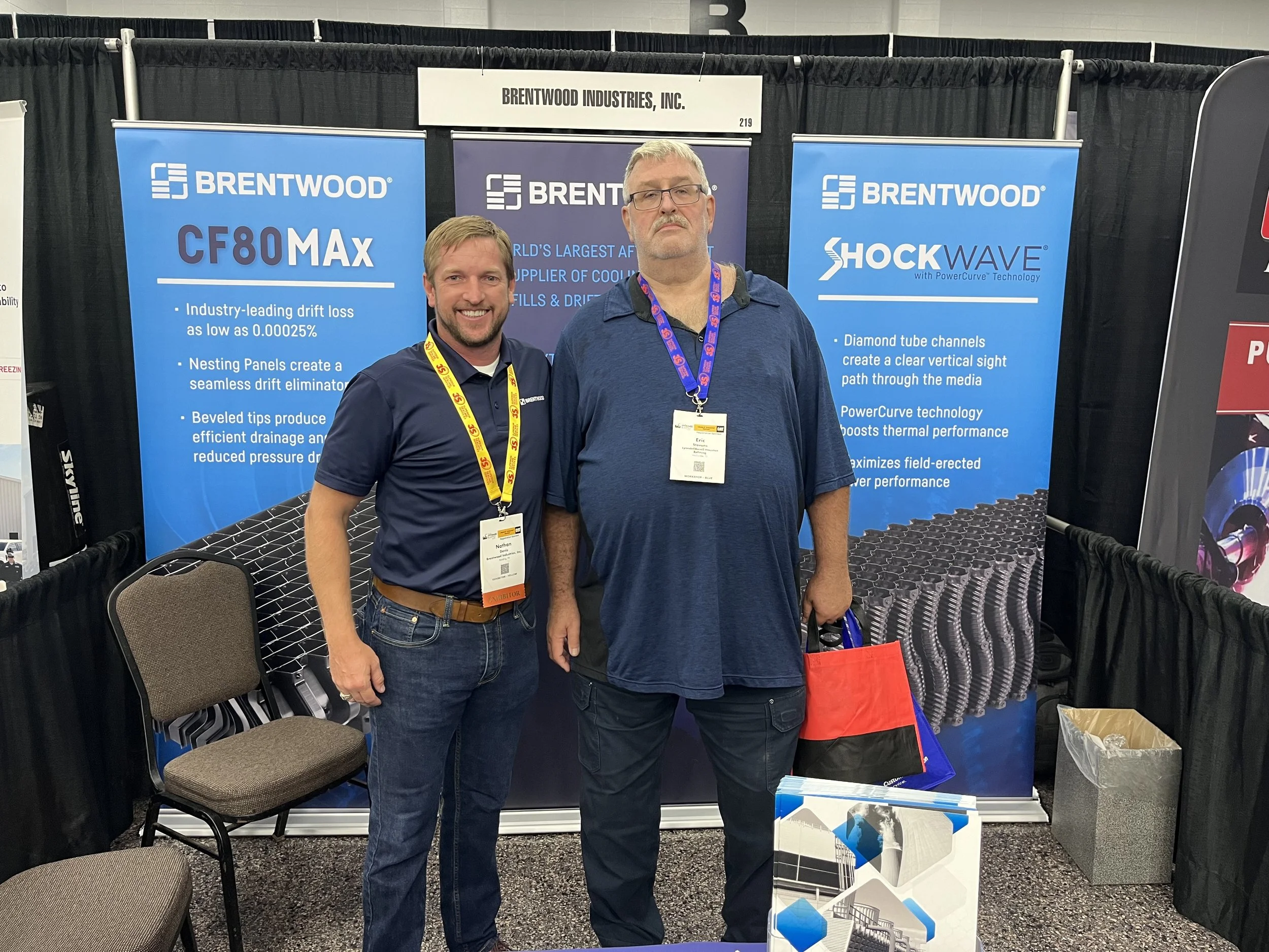 Two men standing in front of a trade show booth displaying banners for Brentwood Industries, Inc., with one man smiling and the other holding a red and black bag.