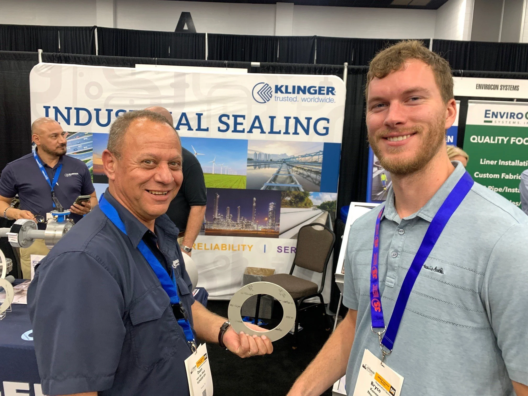 Two men smiling at a trade show booth for Klinger Industrial Sealing, with banners displaying industrial infrastructure and pipelines in the background. One man holds a metal gasket, and both wear conference badges.