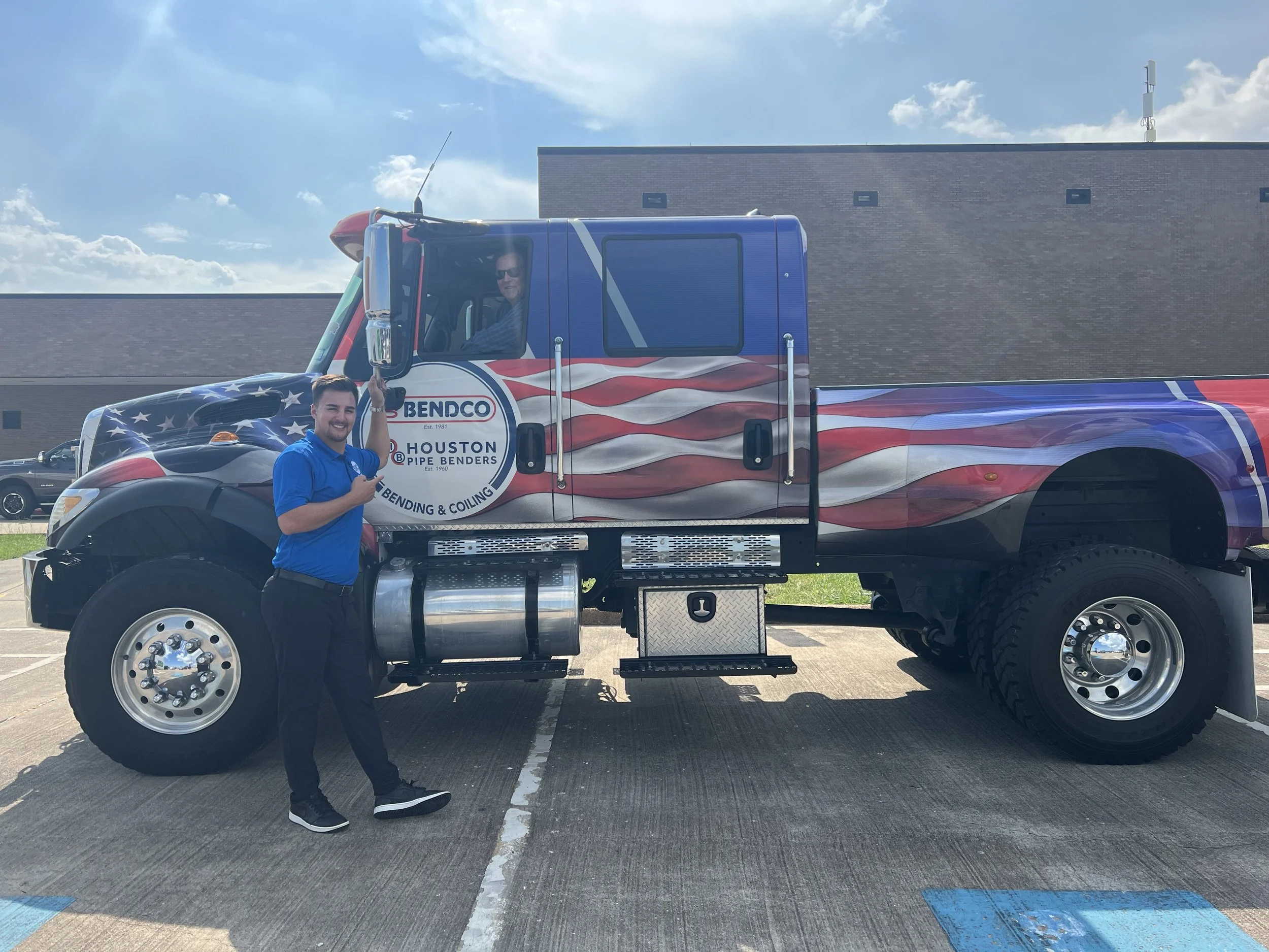 A man in a blue shirt standing next to a large truck with an American flag design, holding keys, with a person inside the driver's seat.