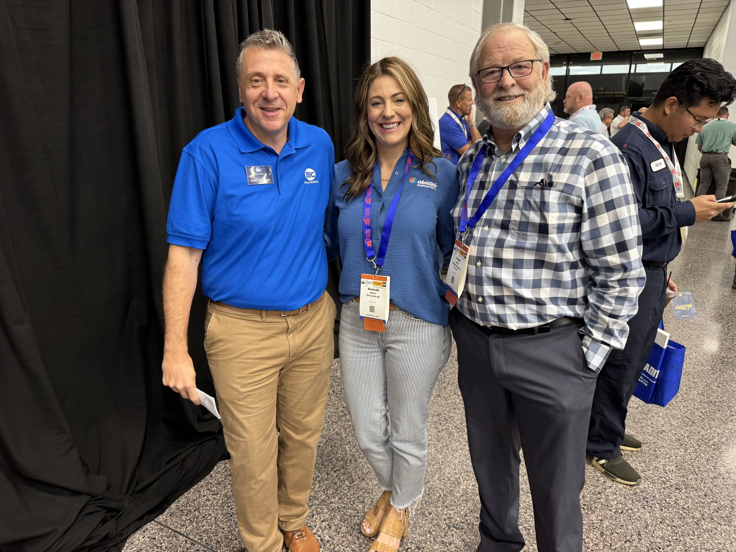 Three people standing together at a conference, smiling for the camera. The person on the left is wearing a blue polo shirt with a name tag, the woman in the middle is wearing a blue top with conference lanyard, and the man on the right is wearing a 