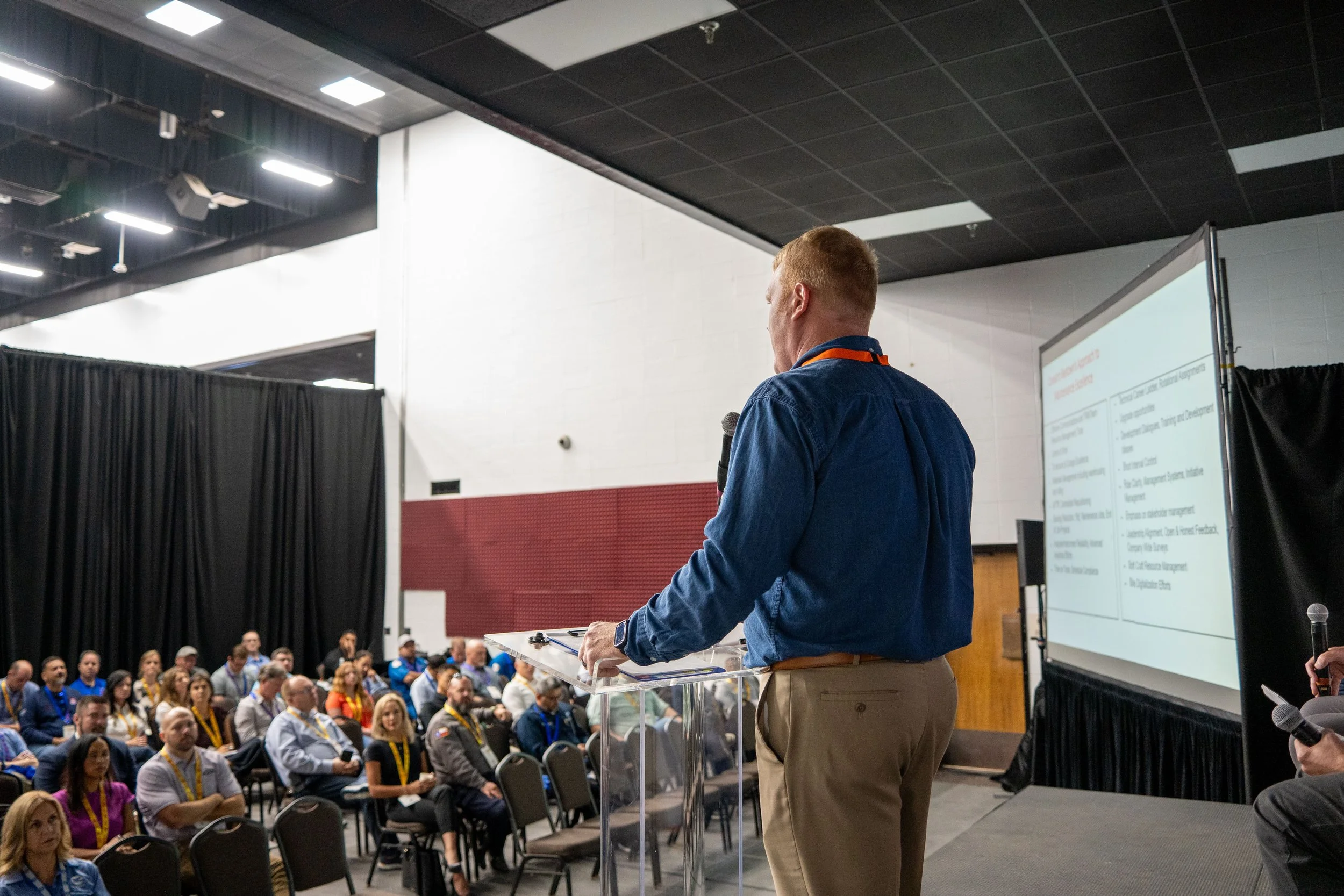 A man in a blue shirt and beige pants speaks into a microphone while standing at a podium in front of an audience. The audience is seated and listening, with some taking notes. There is a large screen displaying a presentation to the right of the spe
