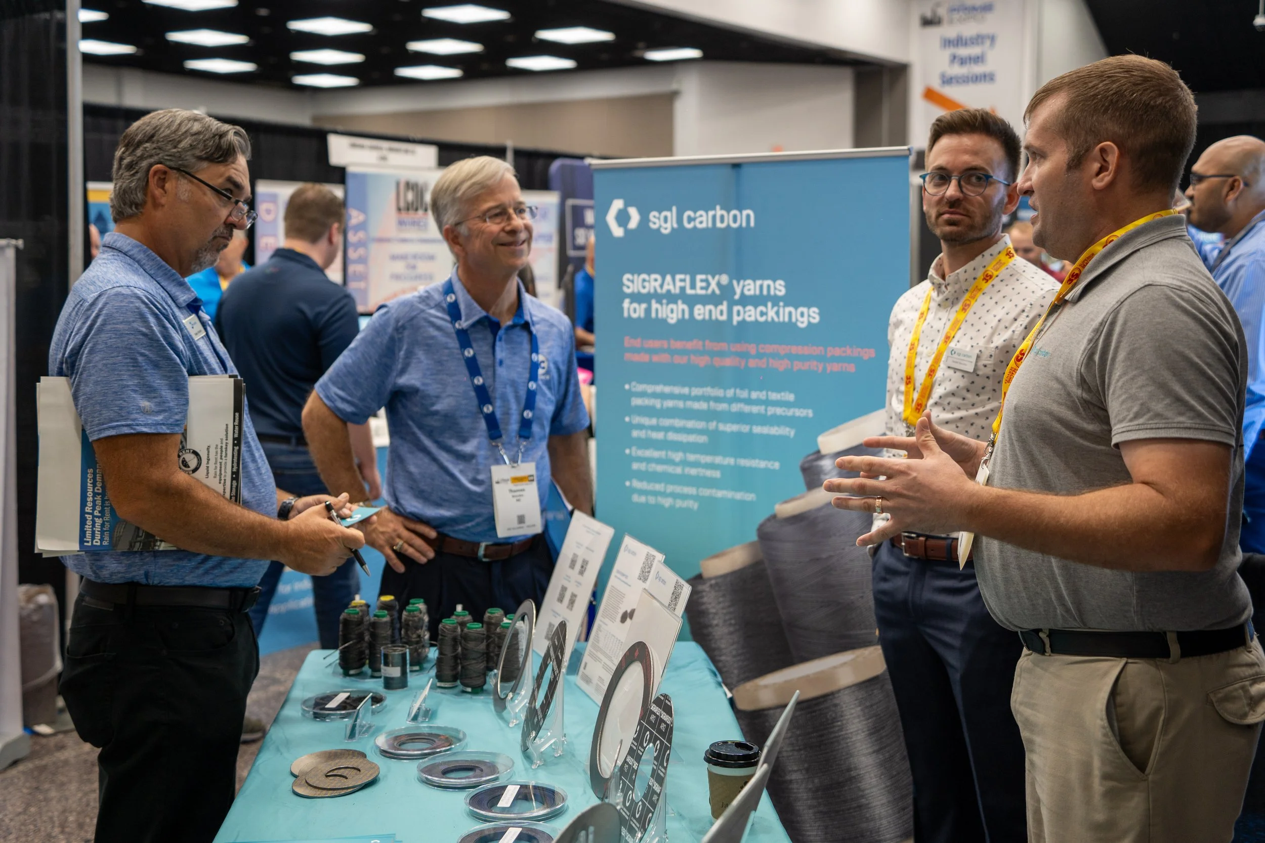 People attending a trade show booth discussing products related to high-end yarns and coatings. The booth features a banner with the text 'sjl carbon' and 'SIGRAFLEX yarns for high end packings.' Various samples and informational materials are displa