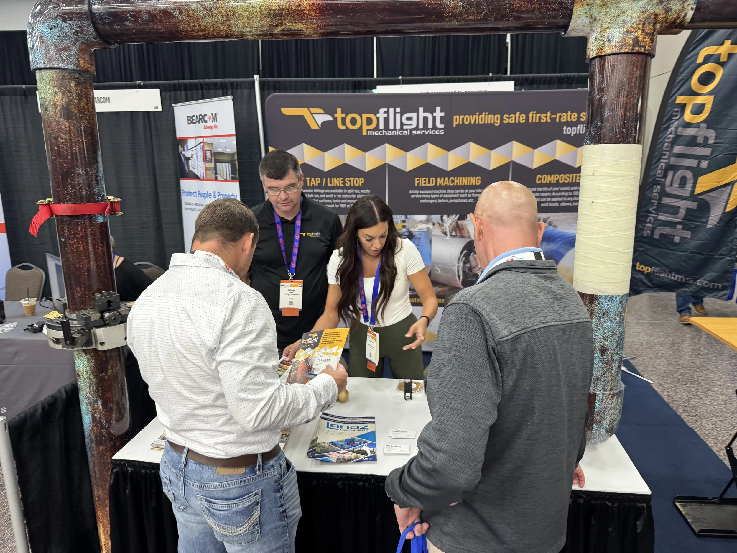 Two men and one woman at a trade show booth discussing with a visitor. The booth promotes a company called 'topflight' offering mechanical services like tape line stop, field machining, and composite work. The background has banners with the company'