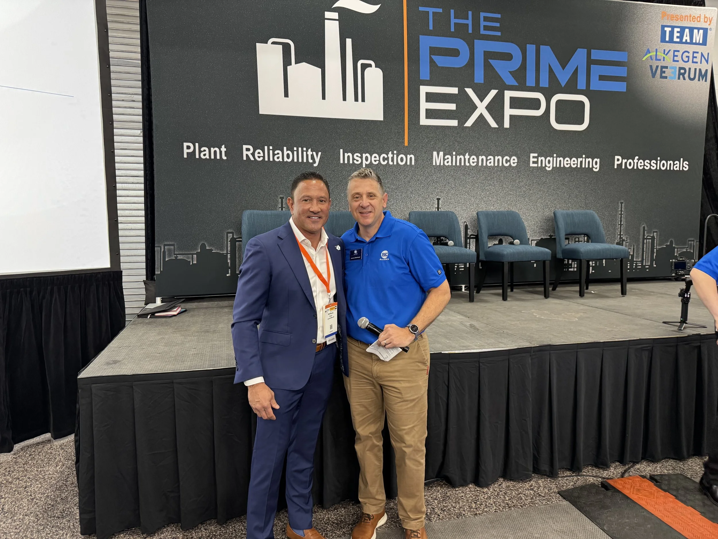Two men standing in front of a stage at The Prime Expo, posing for a photo. The stage backdrop features a large graphic with text about plant reliability, inspection, maintenance, engineering, and professionals. The man on the left is wearing a blue 