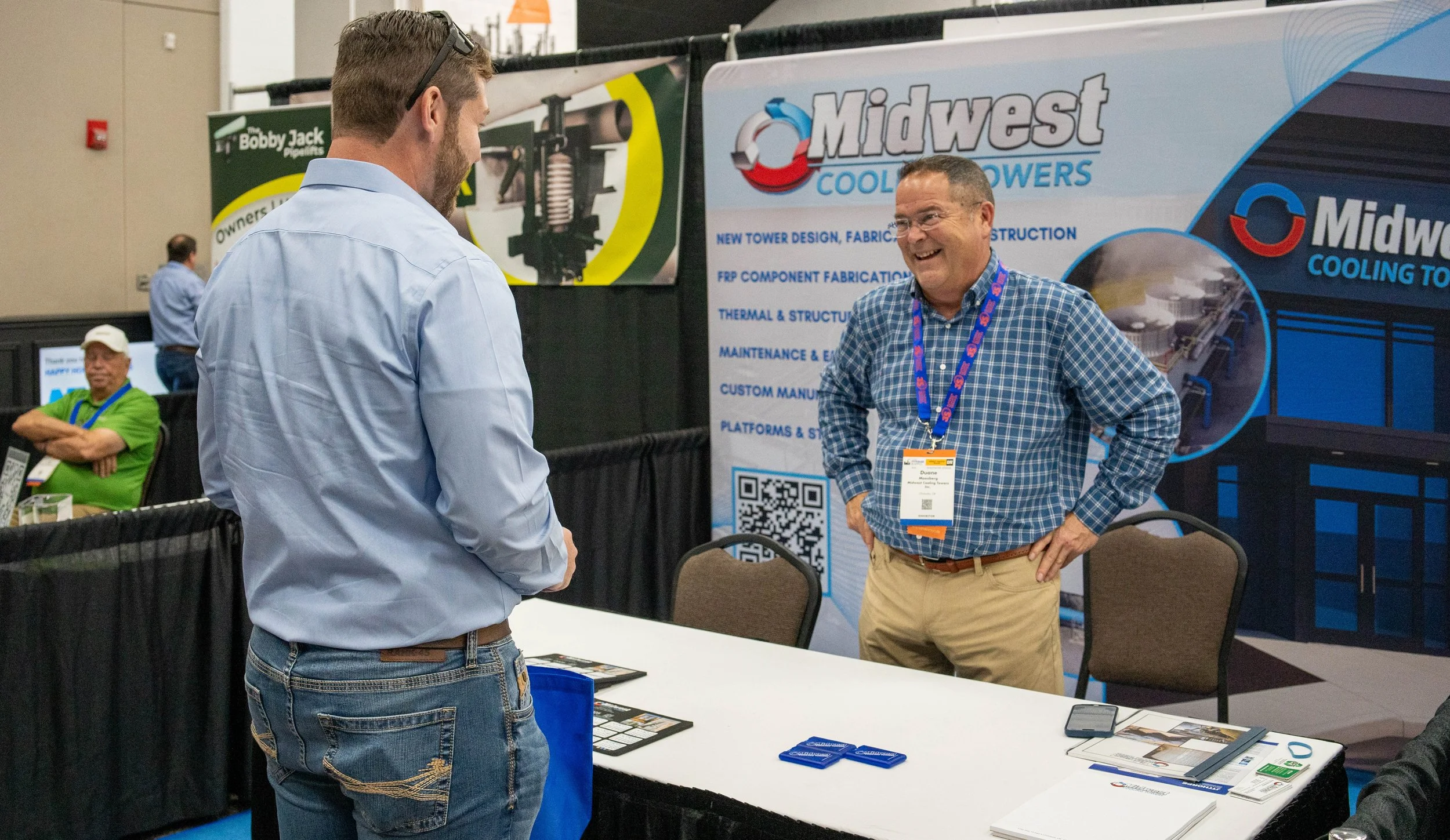 Two men having a conversation at a trade show booth, with one wearing a blue checkered shirt and the other in a light blue shirt. There are banners for Midwest Cooling Towers in the background.