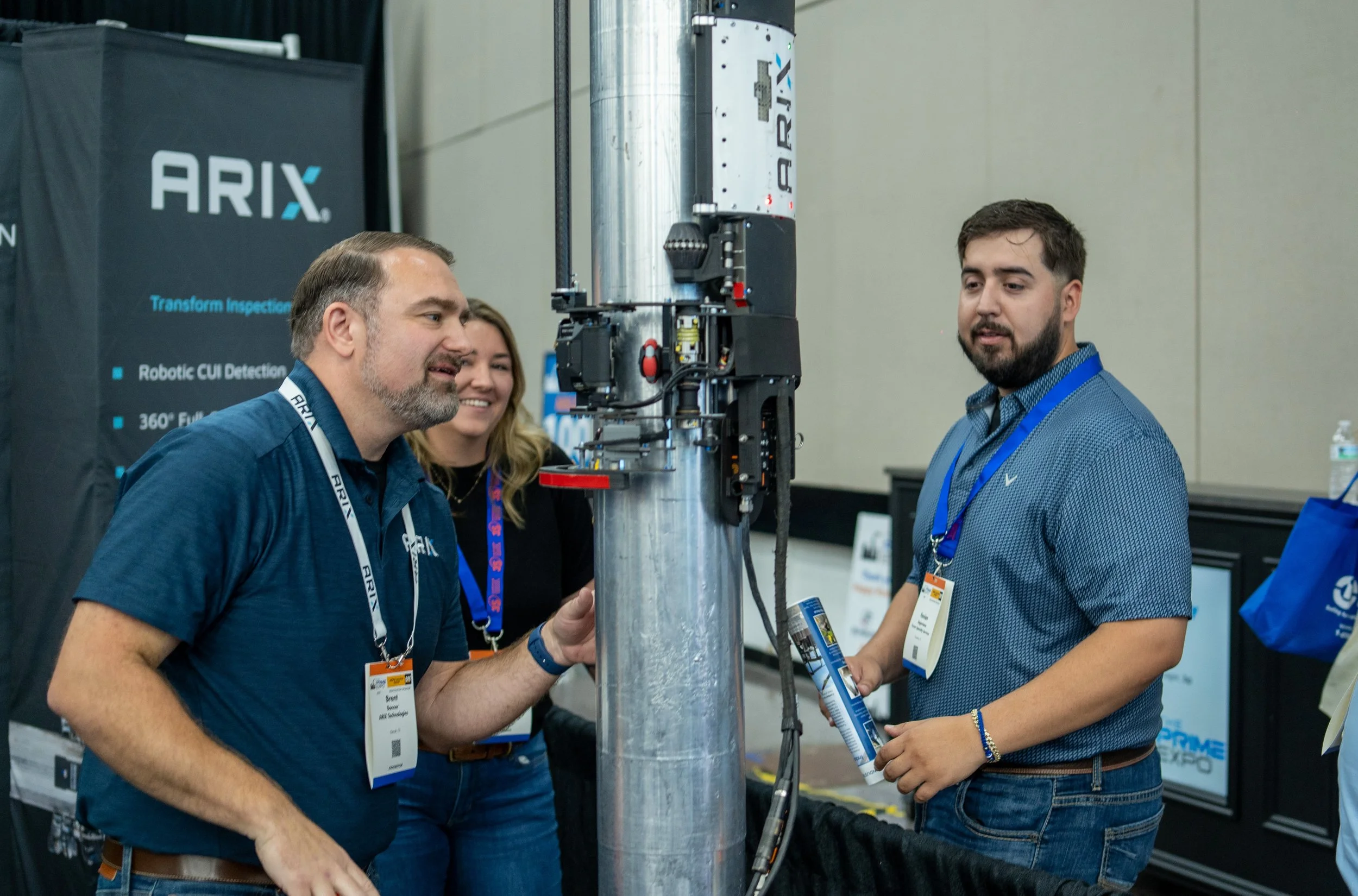 Three people standing around a vertical robotic arm at a technology expo. They are engaging with the robot, which has a sleek metallic body and various sensors and wires. The background includes signage for ARIX with blue and black text and a confere