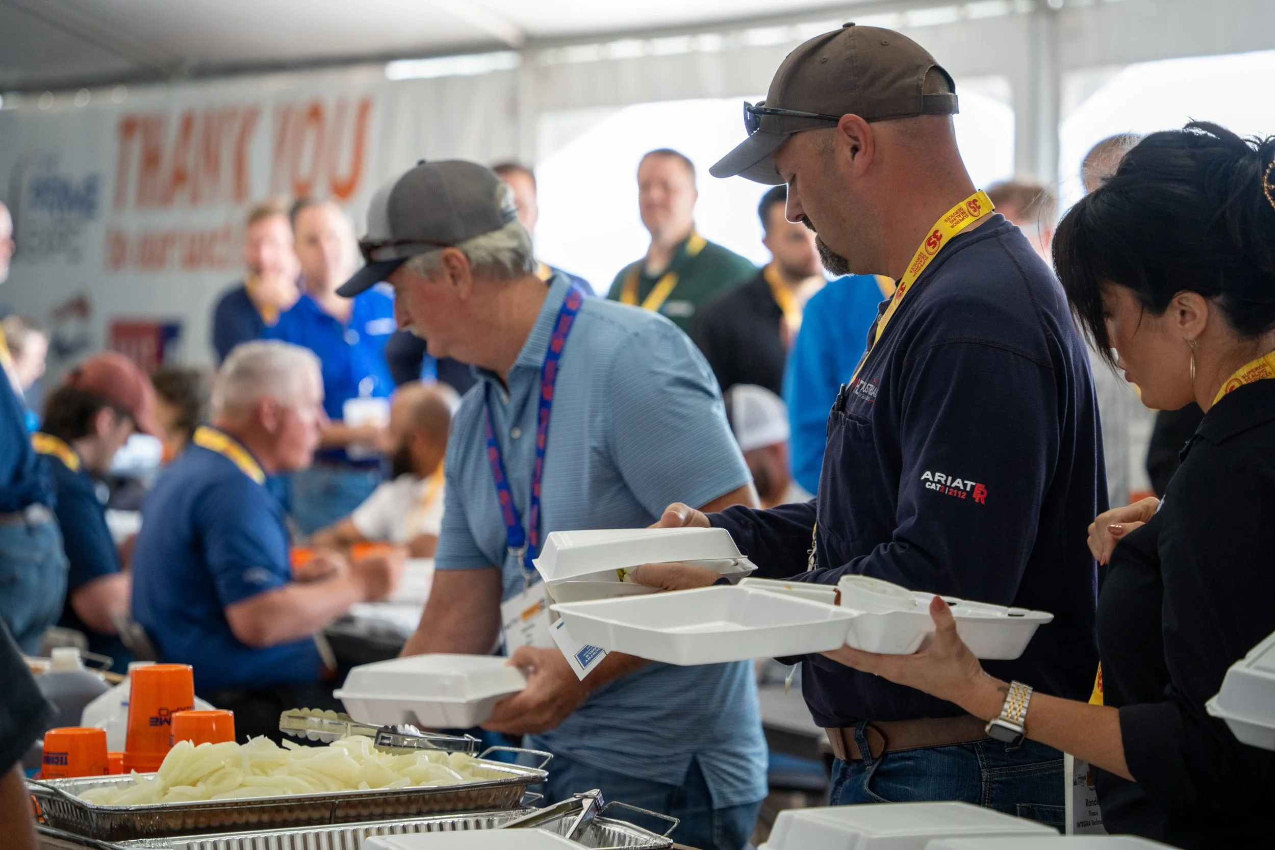 People in a crowded mess hall serving and getting food, with a large banner in the background that says "Thank You" and other text.