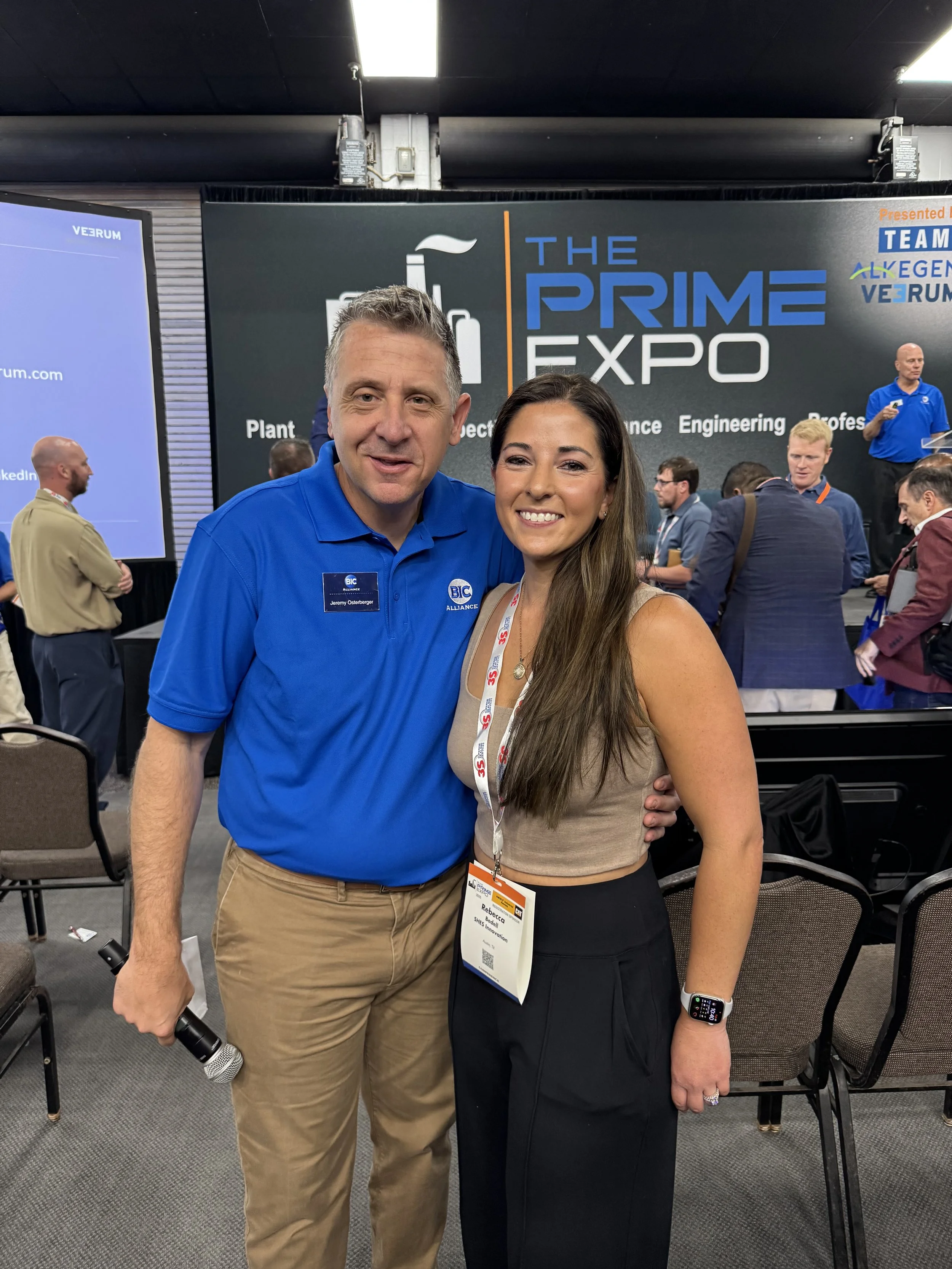 A man and woman smiling and posing together at a conference, with a large sign behind them reading 'The Prime Expo' and other conference attendees in the background.