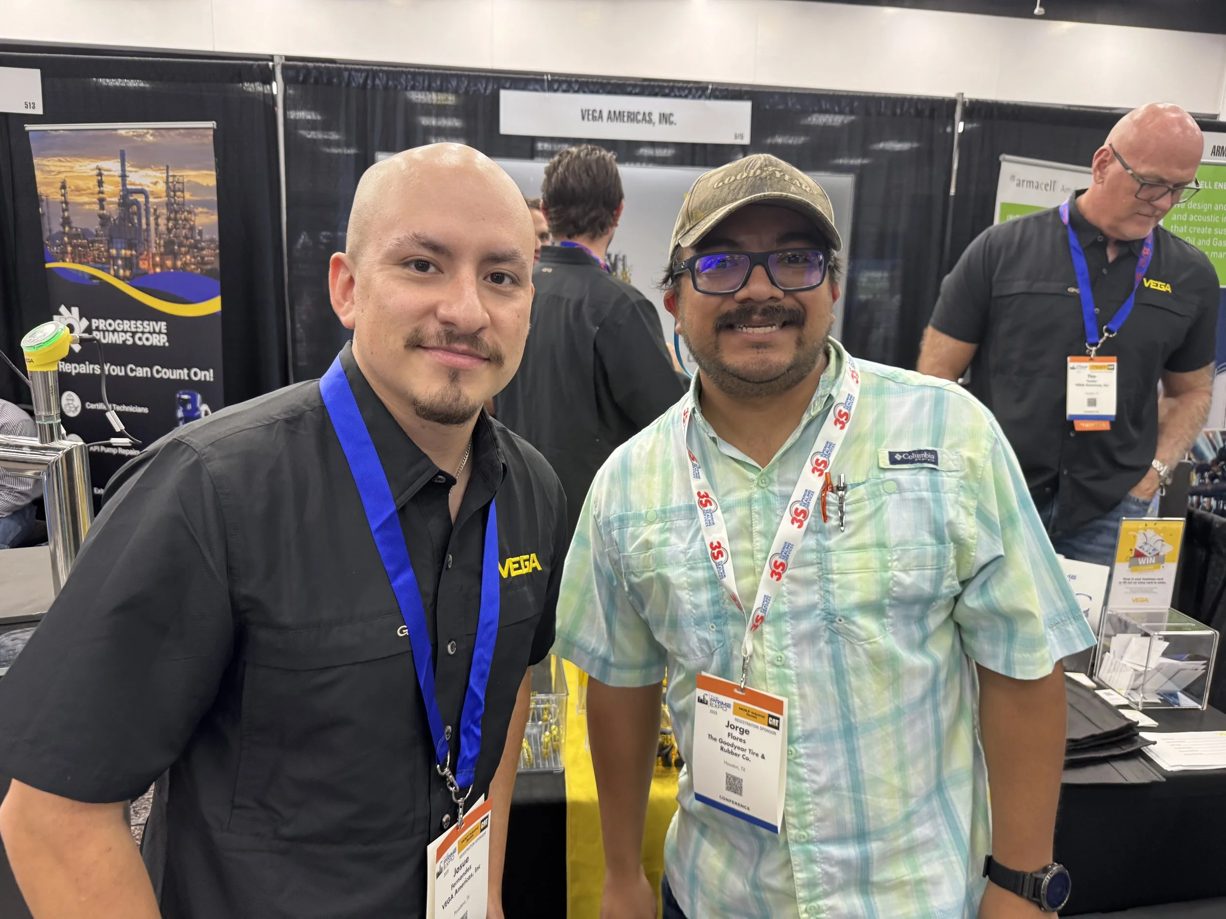 Two men smiling at a trade show booth. The man on the left is wearing a black shirt with a VEGA logo and a blue lanyard. The man on the right is wearing glasses, a green plaid shirt, a cap, and a lanyard with event badges.