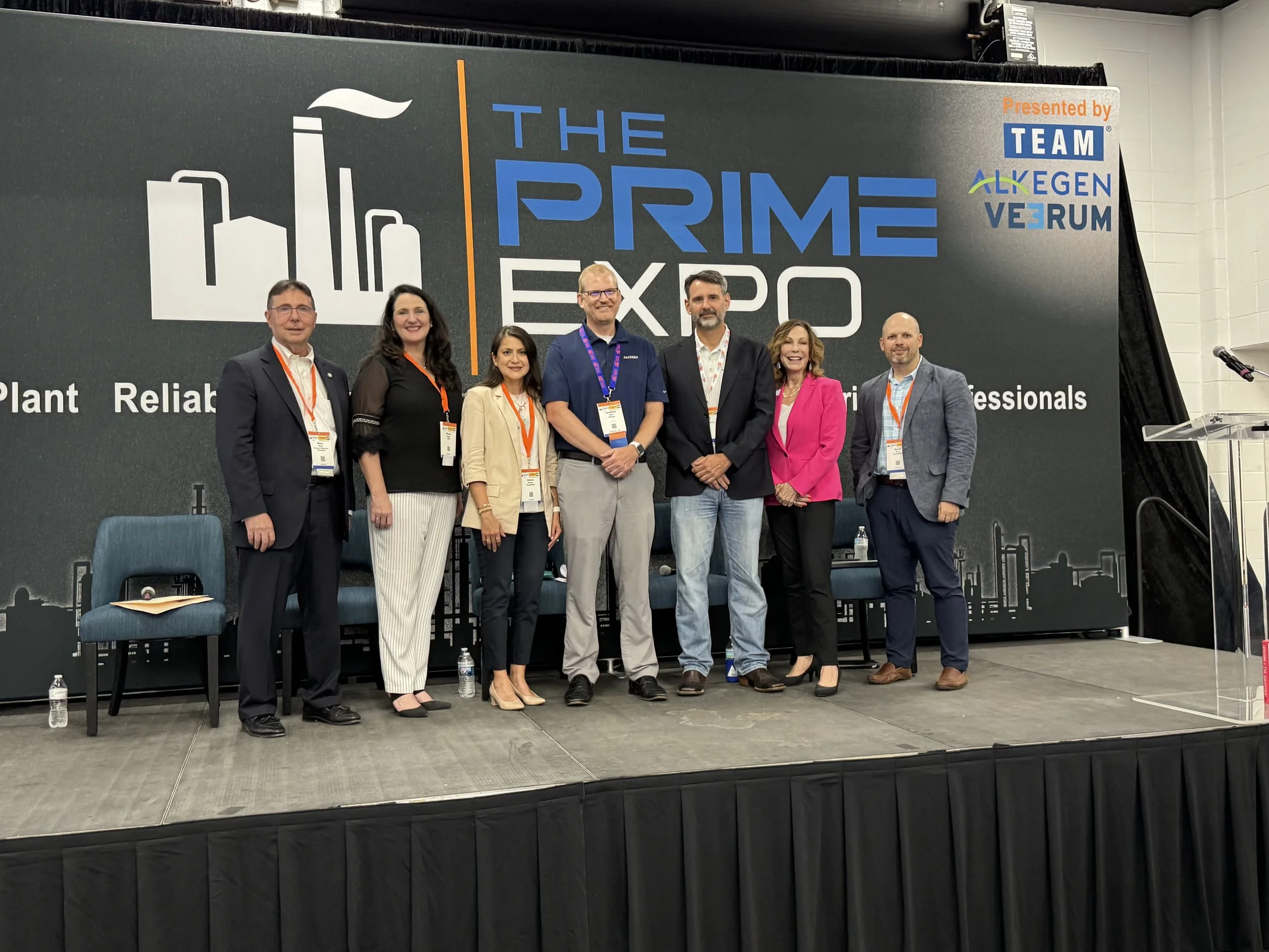 Group of seven people standing on a stage at The Prime Expo conference, with a dark backdrop featuring the event name, logo, and sponsor logos, and a podium to the right.
