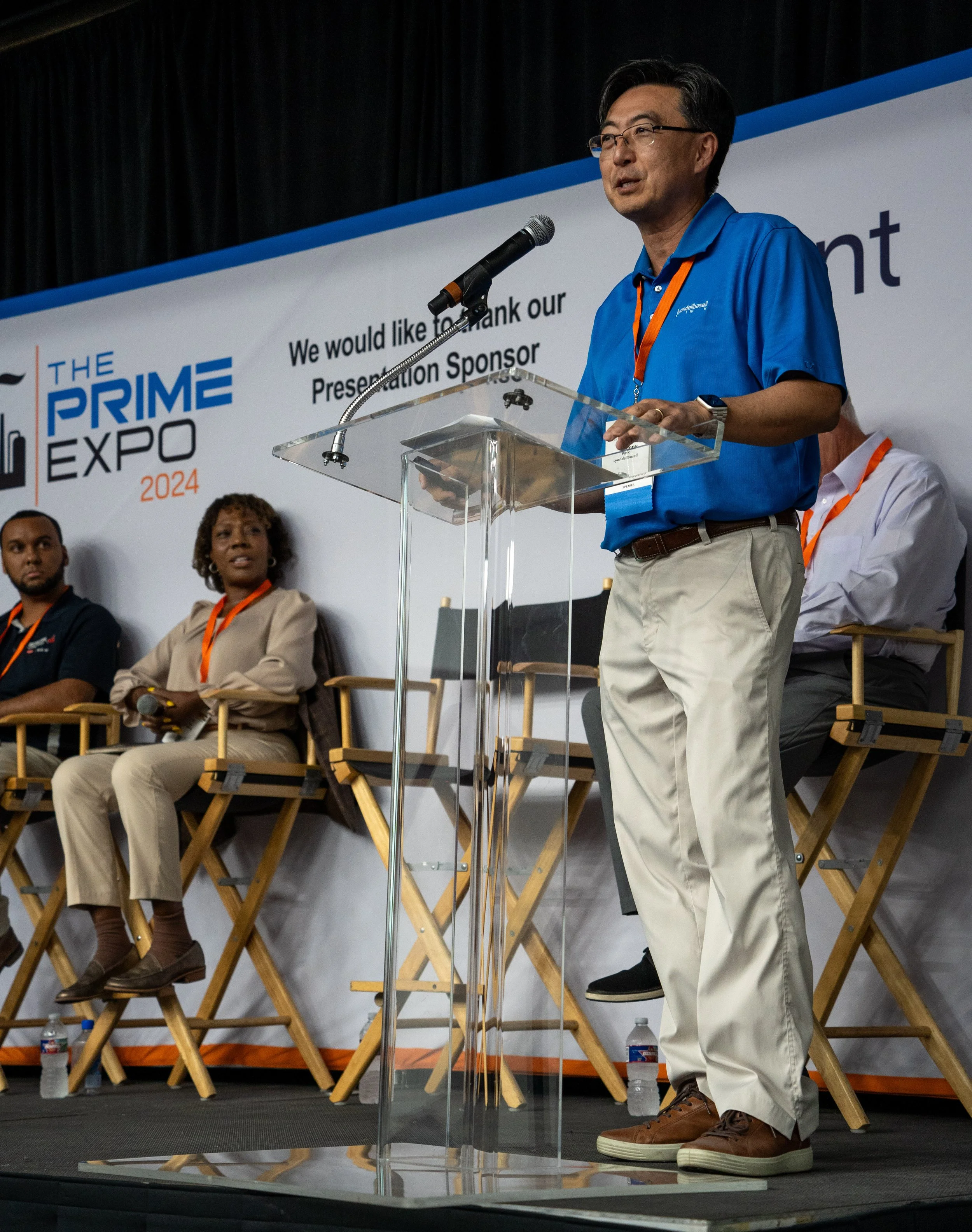 A man in a blue shirt and beige pants stands at a clear podium speaking into a microphone during a panel at the Prime Expo 2024. Three people are seated behind him on a stage with a banner that reads 'The Prime Expo 2024' and a thank you message to a