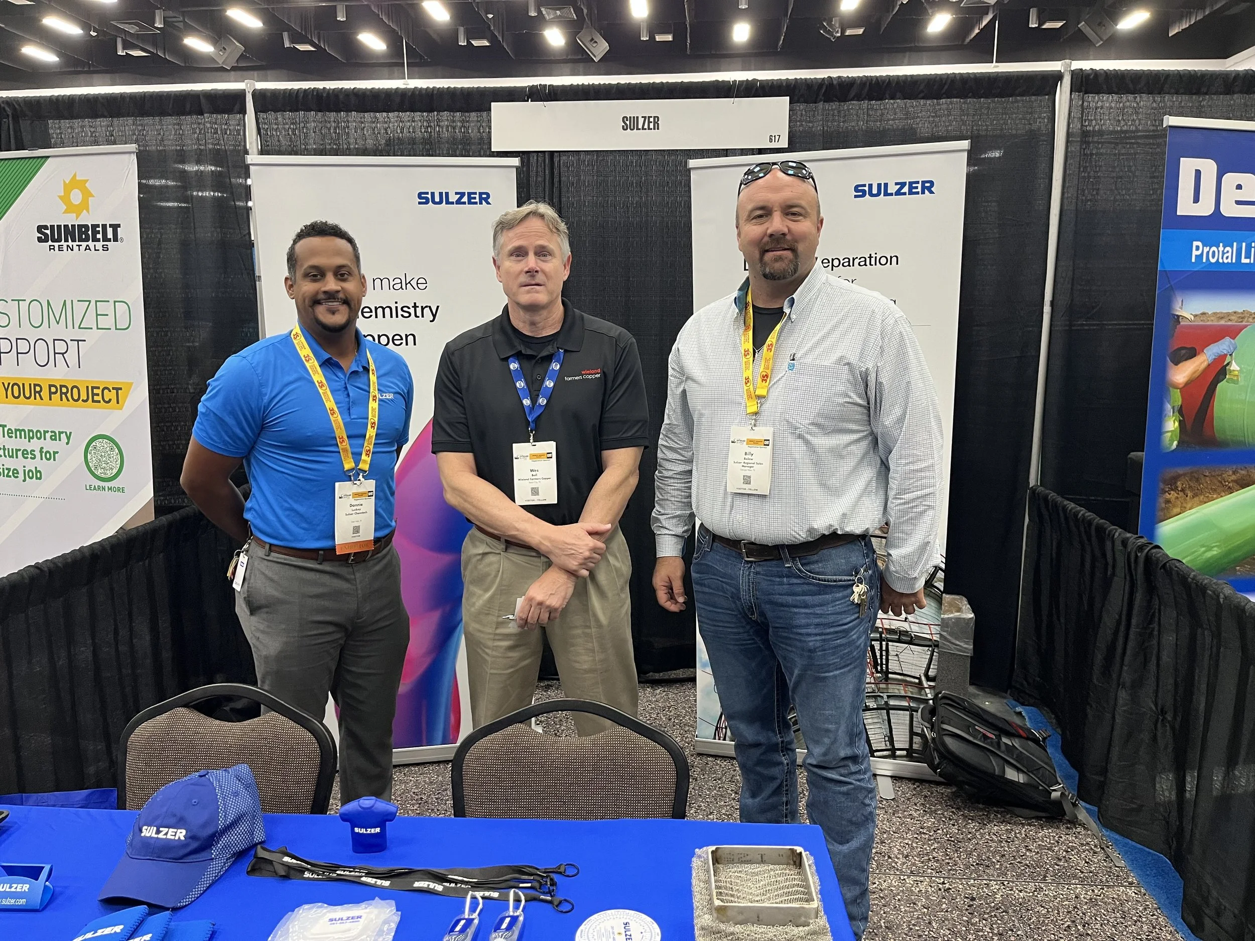 Three men standing at a trade show booth with banners behind them. The booth has a blue table with promotional items, including hats, lanyards, and a small container. The men are wearing name tags and casual business attire.
