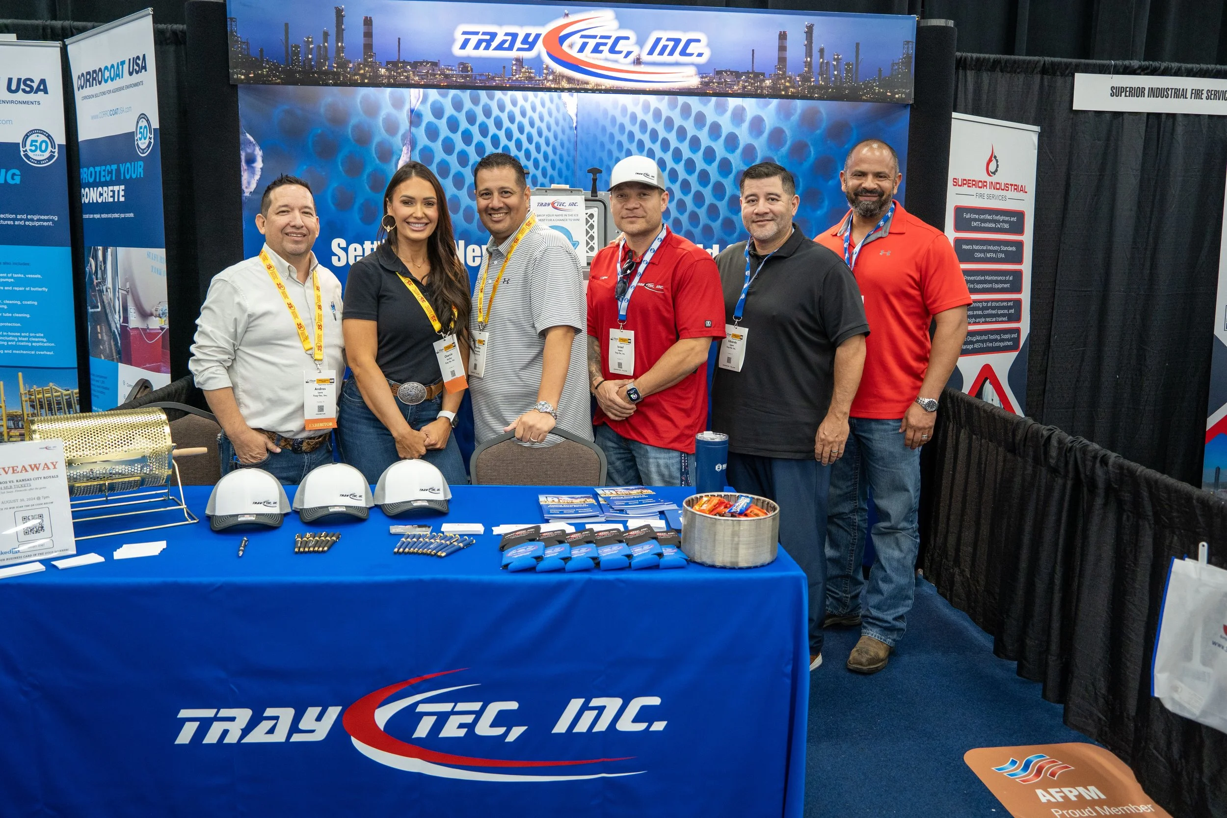 Group of six people standing behind a booth with a blue tablecloth displaying 'Tracy Tec, Inc.' at an industry trade show. The booth has promotional materials, hats, and pens on display, with banners and a city skyline backdrop in the background.