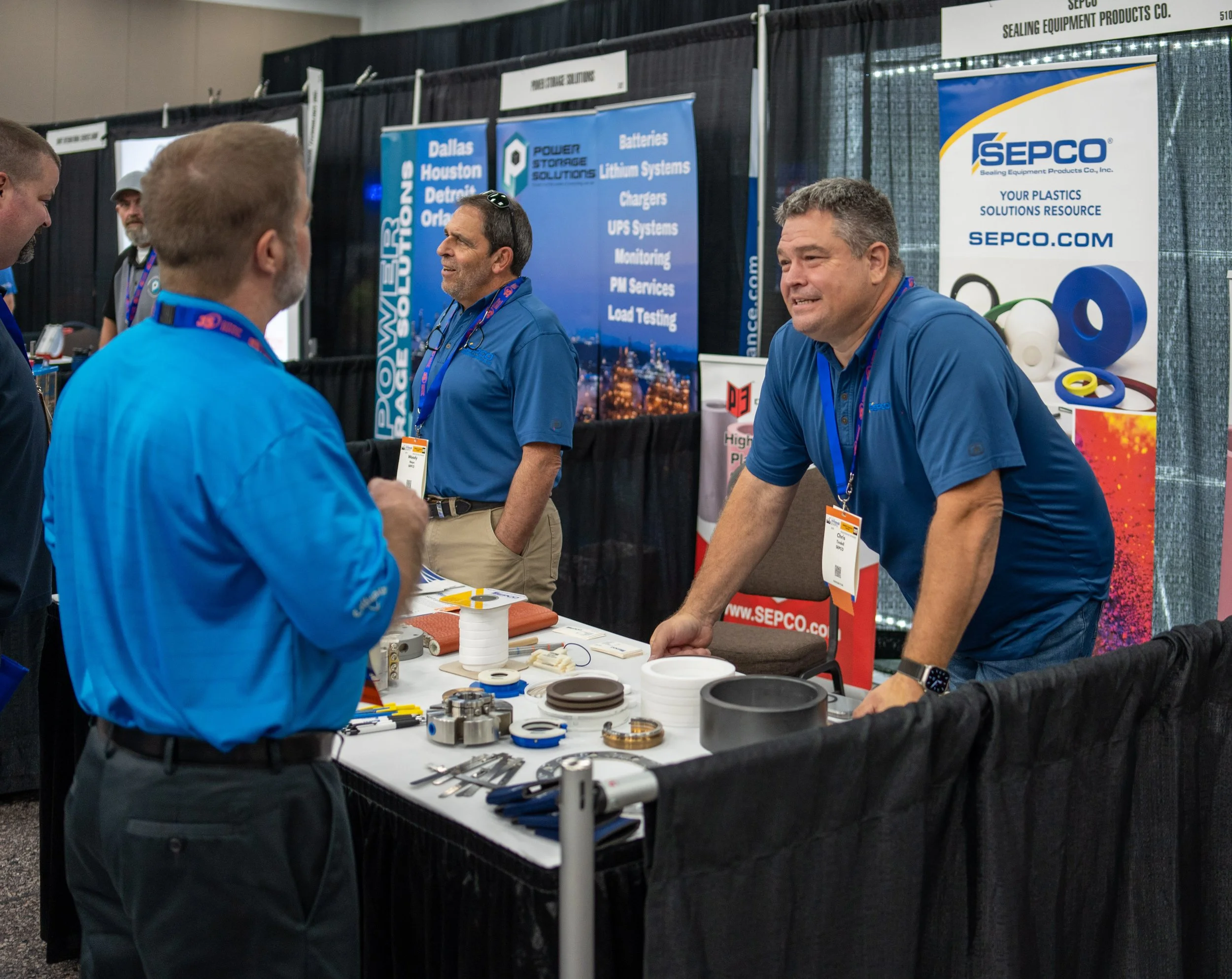 A trade show booth with several men engaging in conversation. The booth displays various industrial parts and equipment, with banners in the background promoting SEPCO and related services. The men are wearing blue shirts and conference badges.