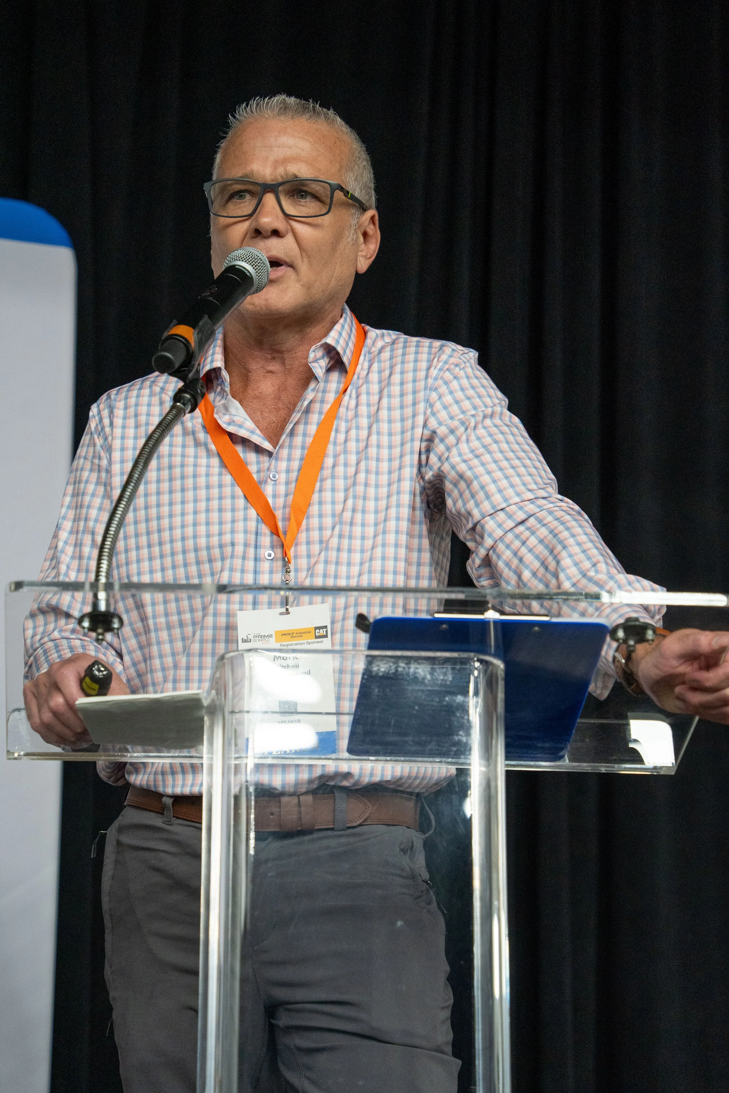 A man speaking at a conference or presentation, standing behind a clear podium with a microphone, wearing glasses, a checkered shirt, and an orange lanyard.
