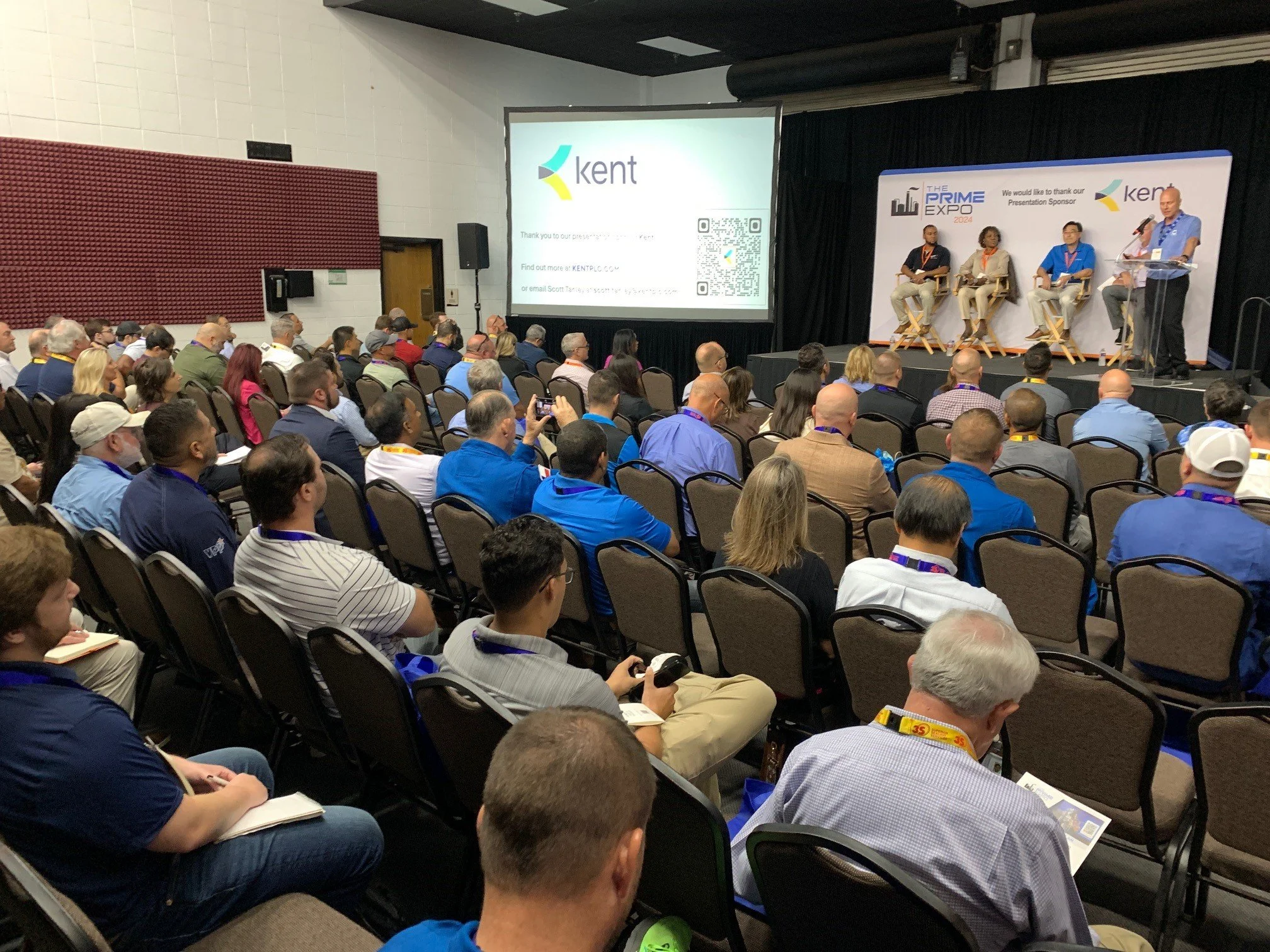 Audience attending a conference with a large screen displaying the Kent logo and QR code, and panelists on stage with a banner in the background reading 'The Prime Expo 2024'.