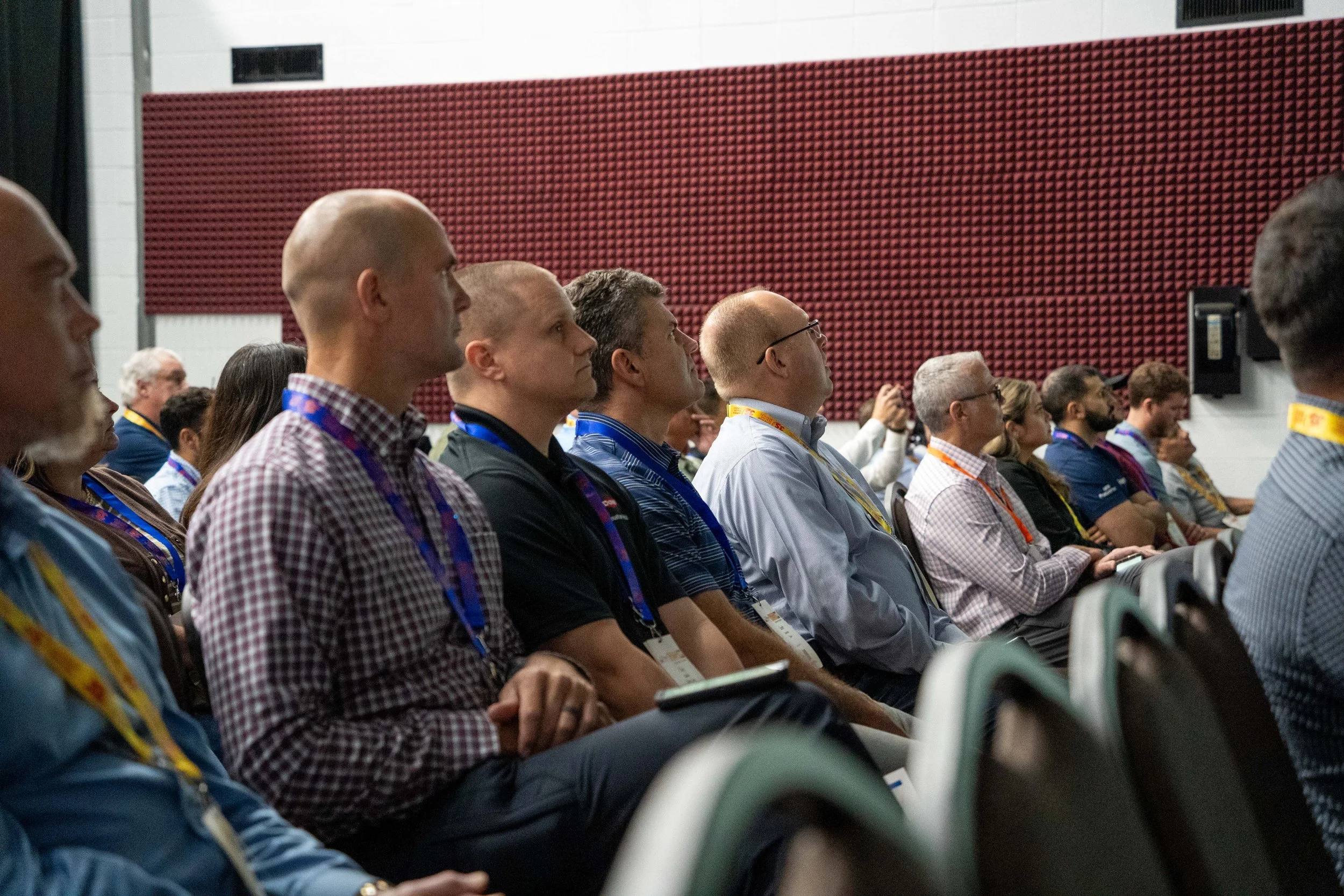 Audience members attending a conference or seminar, seated in rows, listening attentively with a red soundproof wall behind them.