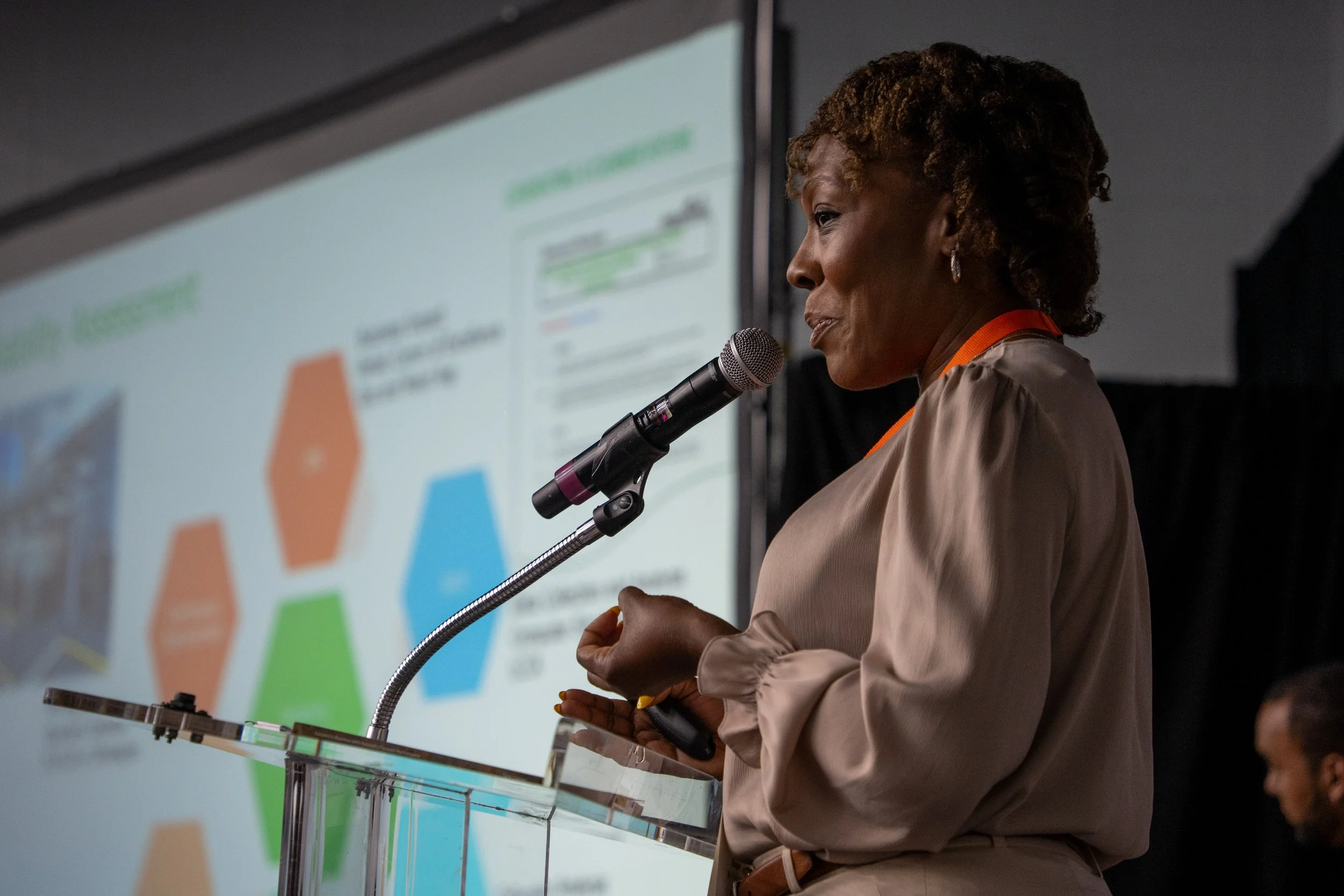 A woman in beige attire speaking into a microphone at a podium during a conference, with a presentation slide featuring colorful hexagons and text displayed behind her.