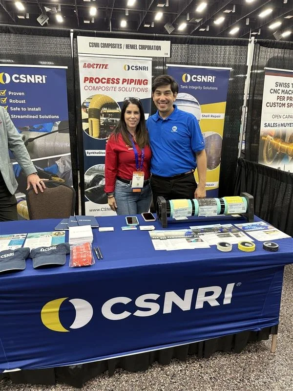 Two people standing behind a booth at a trade show, with banners displaying CSSRI company branding and product offerings such as process piping and tank integrity solutions. The table in front has brochures, pens, and rolls of tape.