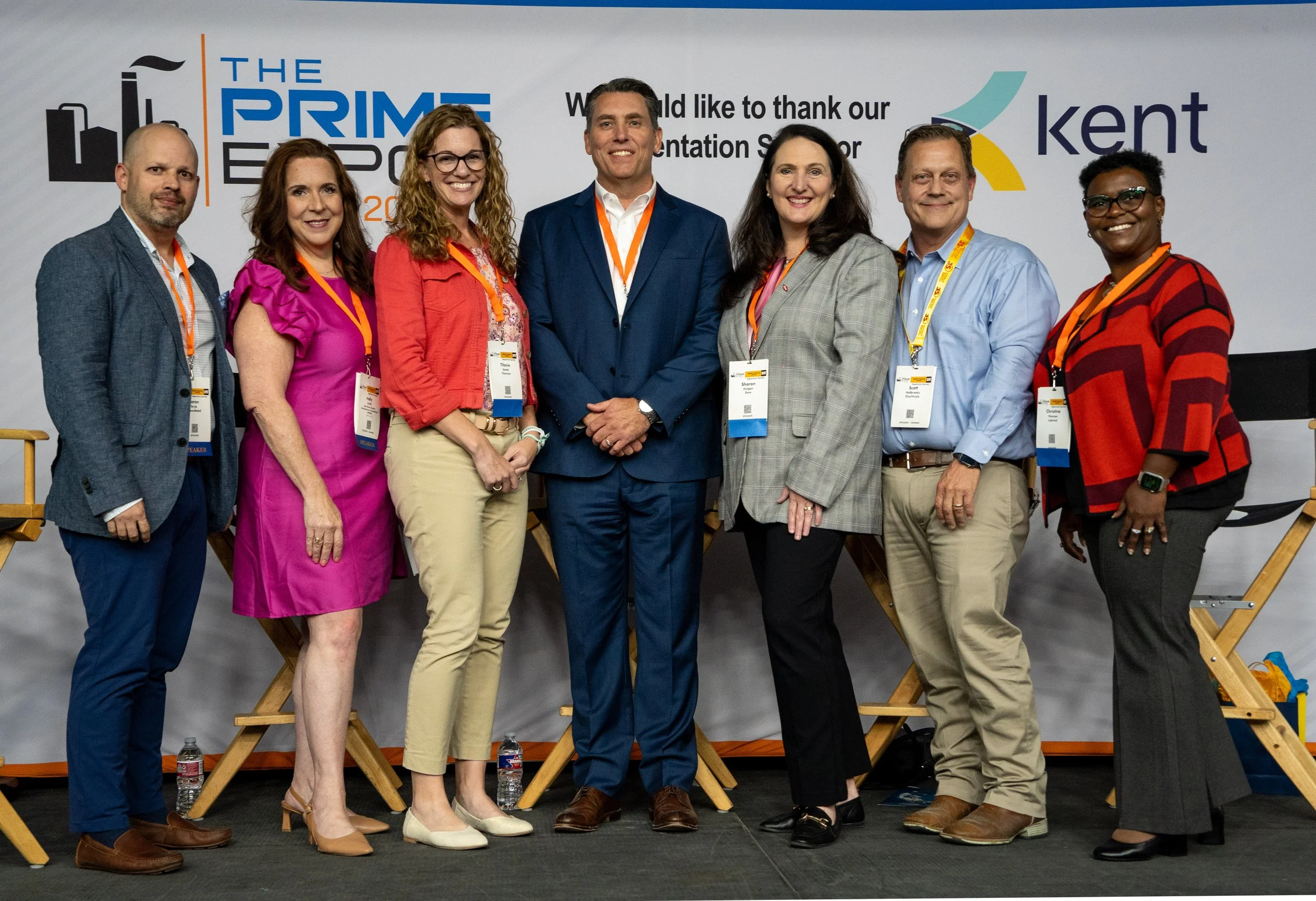 Group of eight diverse professionals standing together at a conference, with banners in the background displaying various logos and the words "The Prime Expo" and "Kent".