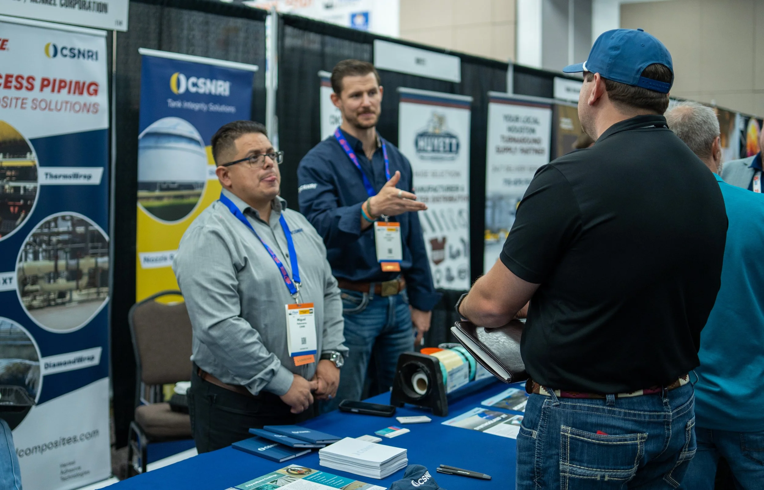 Three men at a trade show booth engaging in conversation. Two men are facing the booth, and one man is talking to them. There are promotional banners and brochures on the table.